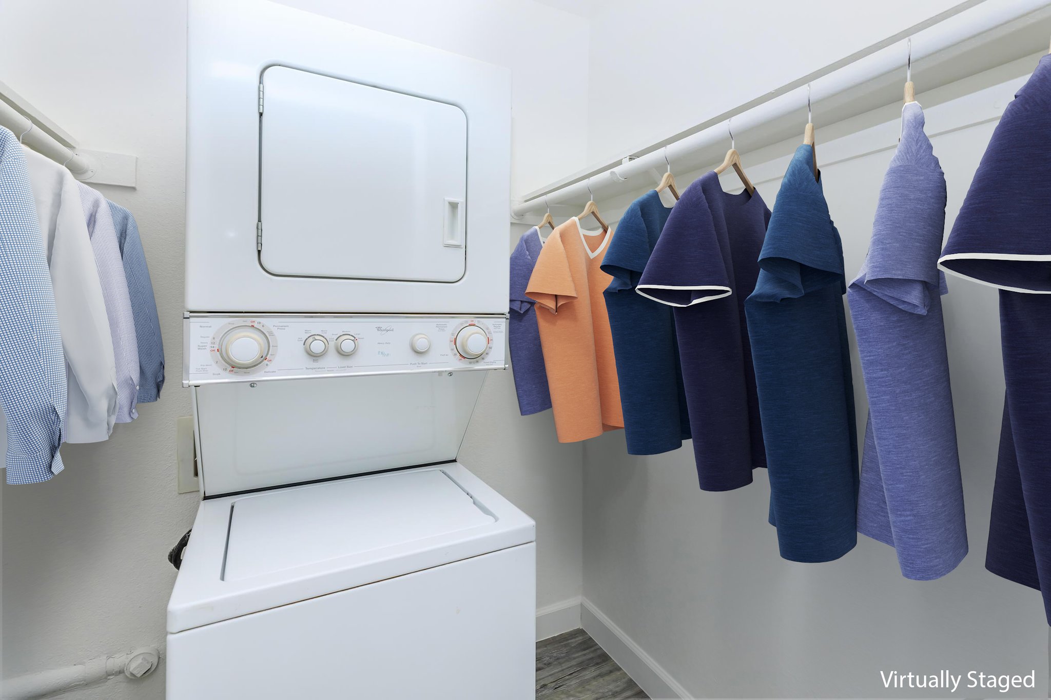 A laundry area featuring a stacked washer and dryer against a wall. To the right, a clothing rack holds various shirts in shades of blue, purple, and orange, arranged neatly. The room has a clean and organized appearance, with light-colored walls and flooring.