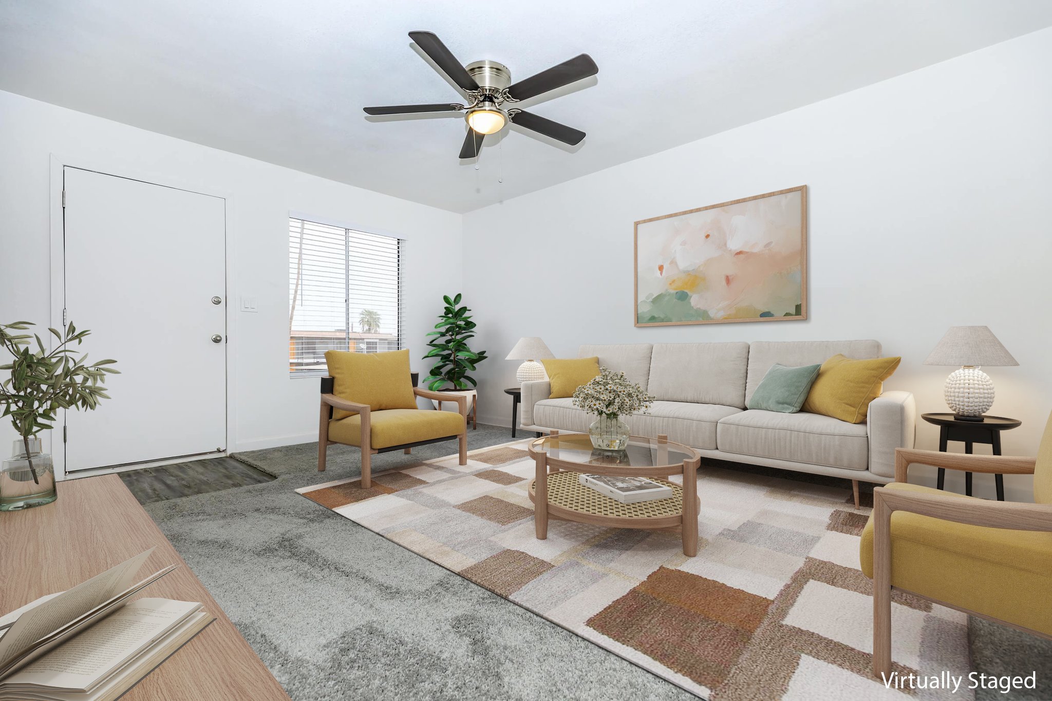 A modern living room featuring a light beige couch with yellow accent pillows, two yellow chairs, and a round coffee table. A large rug with a geometric pattern covers the floor. A potted plant adds greenery, while a ceiling fan provides overhead cooling. A wall art piece hangs above the couch, and a window lets in natural light.