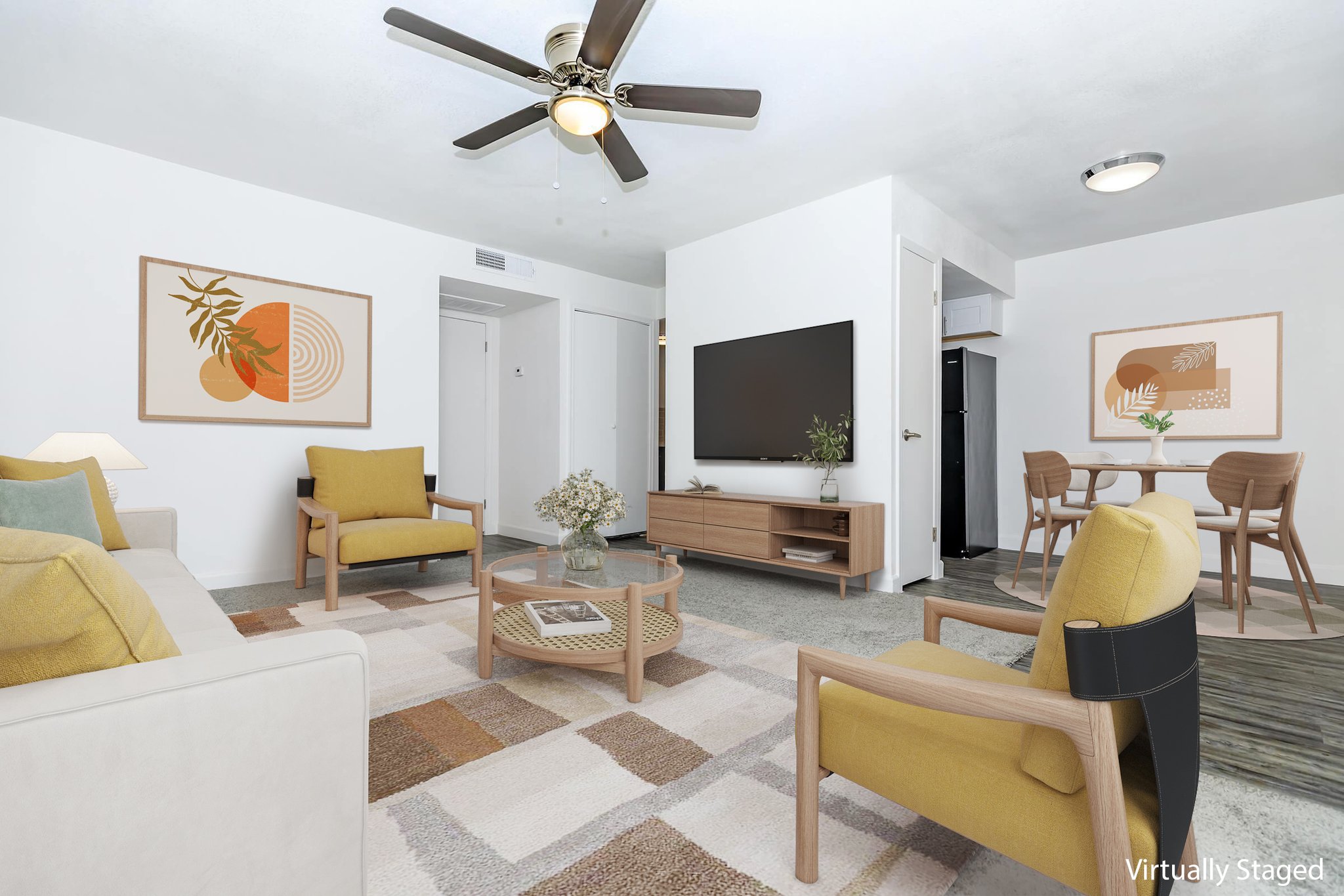 A modern living room featuring yellow and black accent chairs, a wooden coffee table, and a TV mounted on the wall. The decor includes framed abstract art pieces and potted plants. A dining area with a round table and chairs is visible in the background, along with light-colored walls and a ceiling fan.