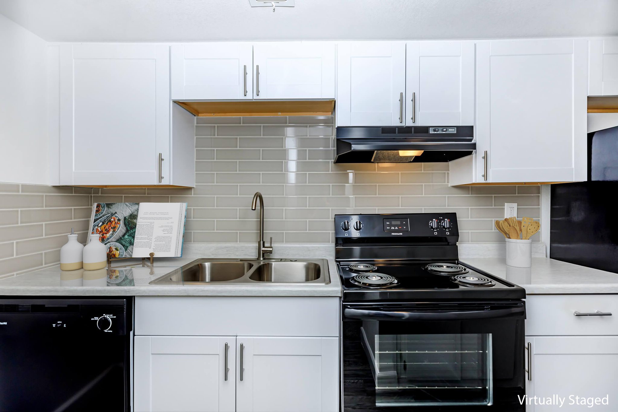 A modern kitchen featuring white cabinetry, a double sink, and black appliances including a stove and dishwasher. The backsplash is made of light gray tiles. A cookbook and decorative items are arranged on the countertop, contributing to a clean and stylish design.