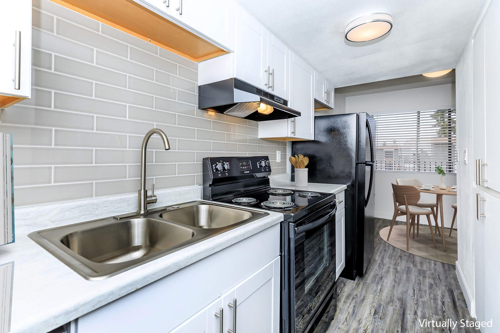 Modern kitchen featuring stainless steel appliances, including a double sink, stove, and refrigerator. The white cabinetry contrasts with a light gray backsplash. Natural light filters through a window overlooking a small dining area with a table and chairs. Clean and contemporary design with ample storage space.