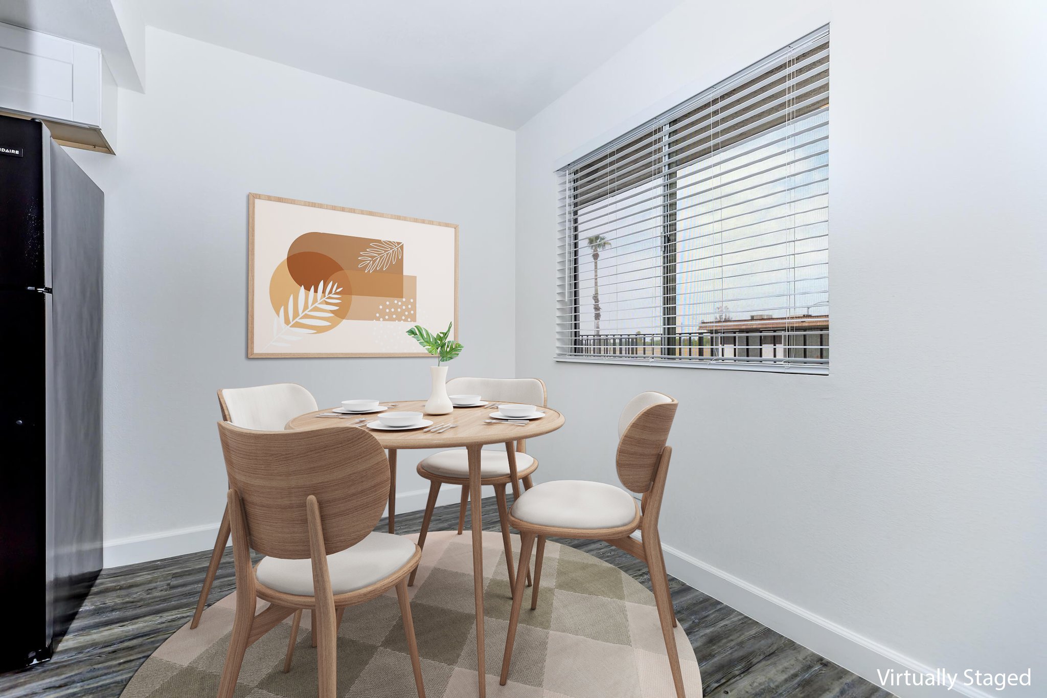 A modern dining area featuring a round wooden table with four white chairs, set with plates and cutlery. A large artwork with abstract shapes in warm tones is mounted on the wall. Natural light filters through a window with blinds, and a small plant centerpiece adds a touch of greenery.