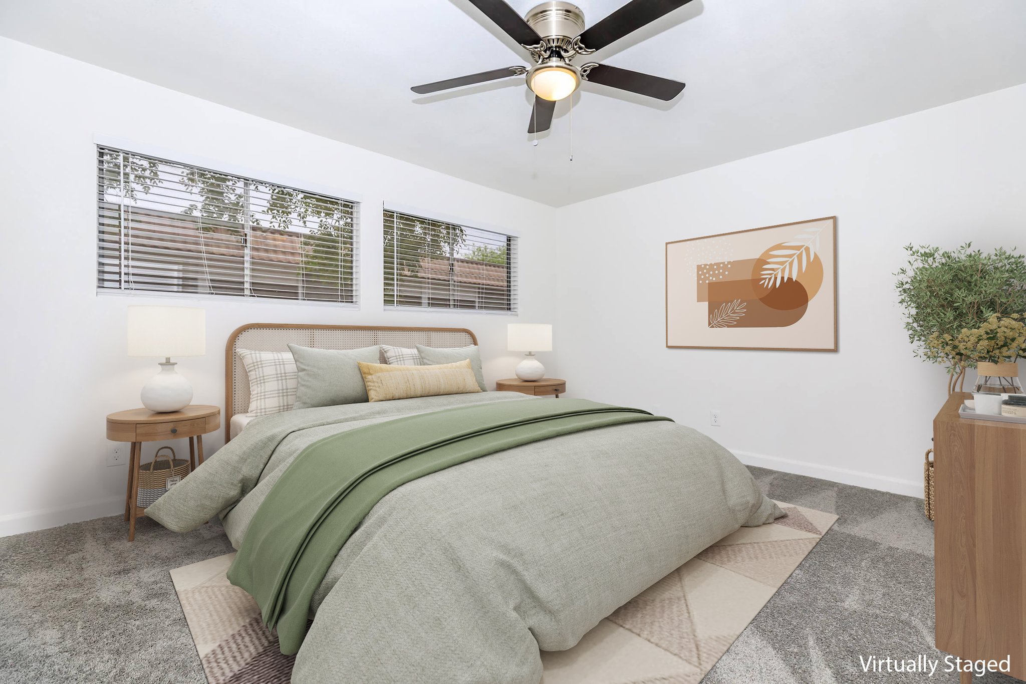 A tastefully staged bedroom featuring a large bed with a green and gray comforter, flanked by round side tables with lamps. There are two windows allowing natural light, a neutral-colored wall art piece, and a small potted plant. The floor is covered with light gray carpet.