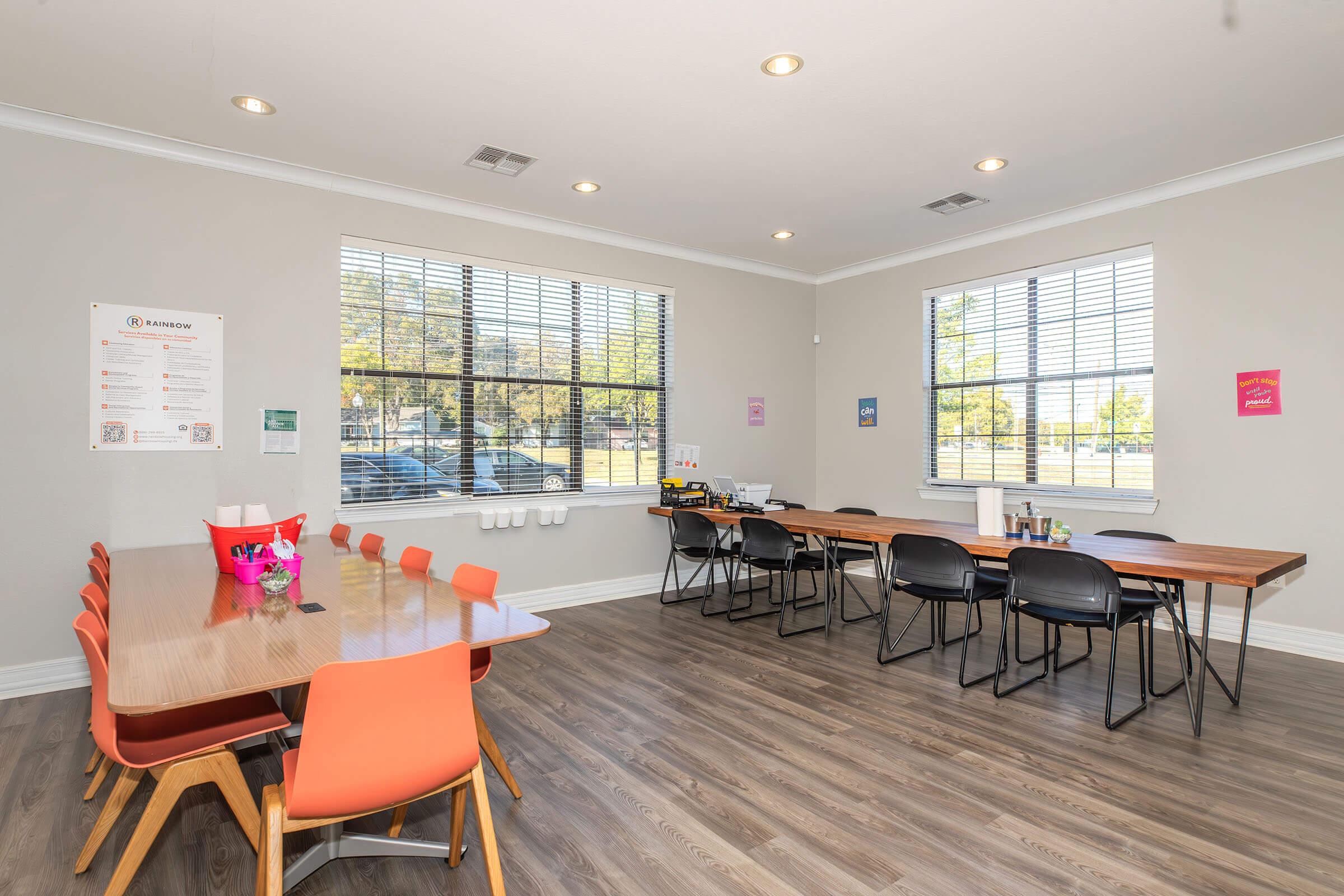 A bright, airy meeting room featuring large windows with blinds, two long wooden tables, and colorful chairs. One table has a pink flower centerpiece and various office supplies. Posters and signs are on the walls, and the floor is a light wood finish, creating a welcoming workspace.