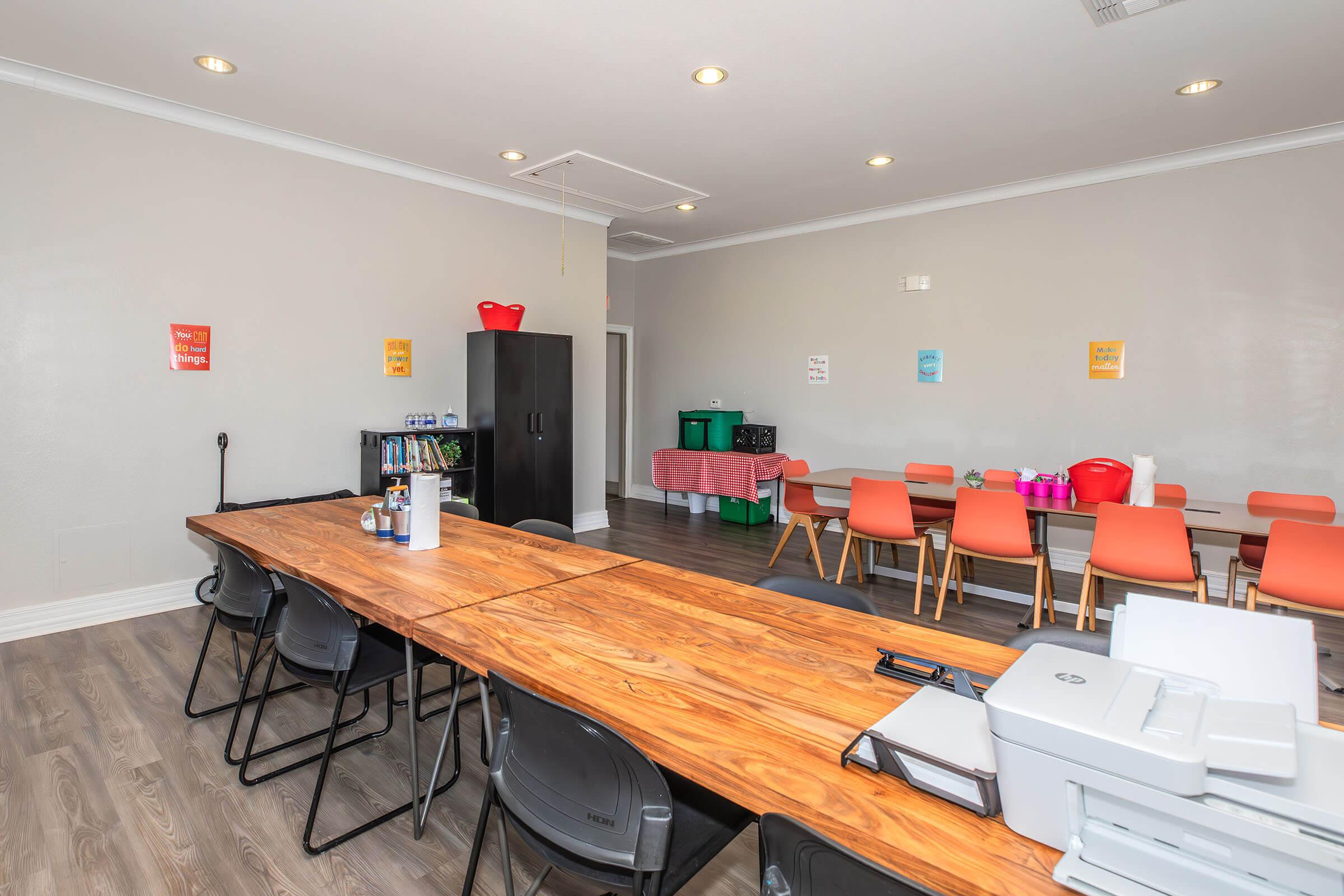 A spacious meeting room featuring a long wooden table with black chairs, a printer, and a storage cabinet. Brightly colored posters adorn the walls, and there are colorful containers on a side table. The room has a modern design with neutral-colored walls and ample lighting.