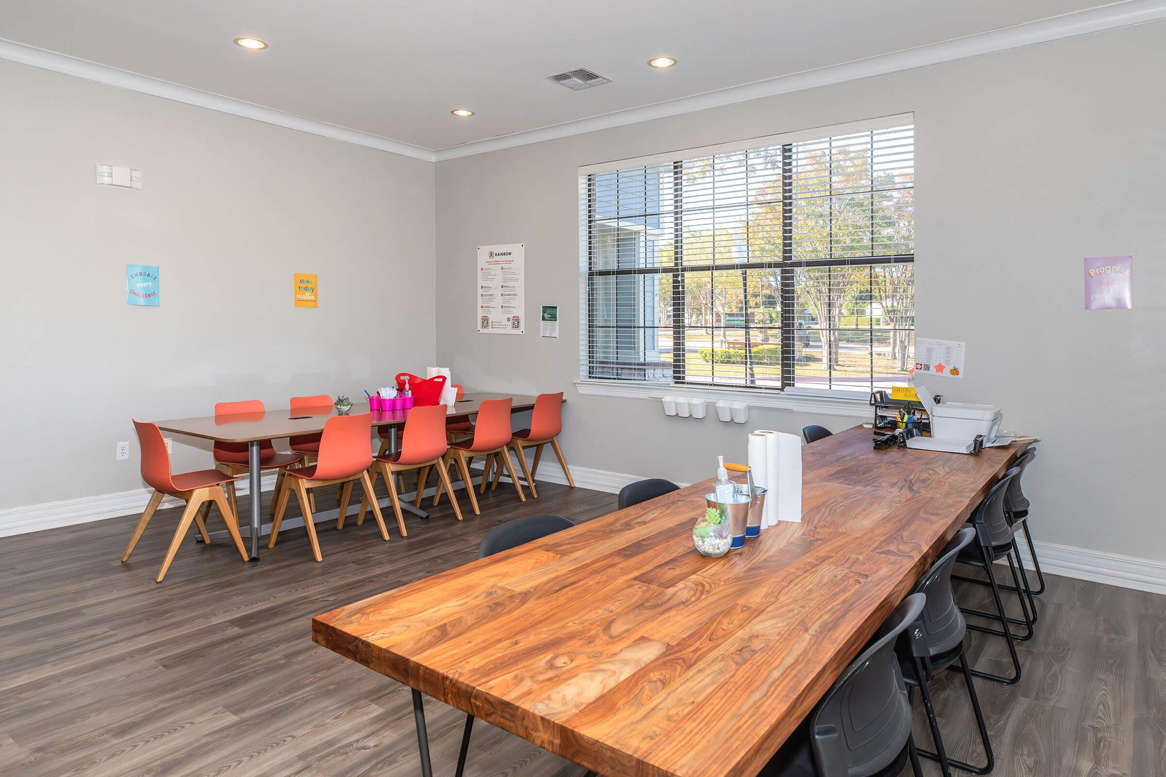 A bright, modern dining area featuring a large wooden table with black chairs and a smaller table with orange chairs. Windows provide natural light, and colorful posters adorn the walls. A serving station with drinks and snacks is visible, creating a welcoming atmosphere.