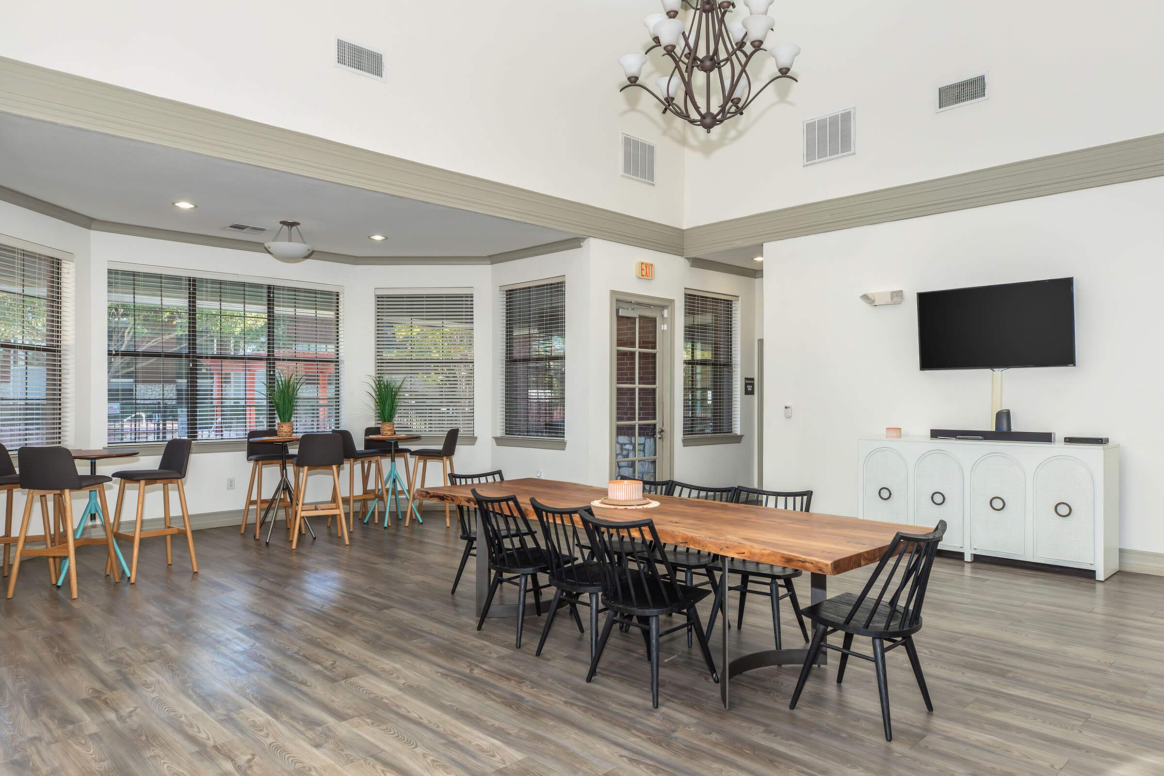 Bright and modern community space featuring a wooden dining table surrounded by black chairs, high-top tables along the walls, and large windows with blinds. A chandelier hangs from the ceiling, and a television is mounted on the wall, complementing the contemporary decor.
