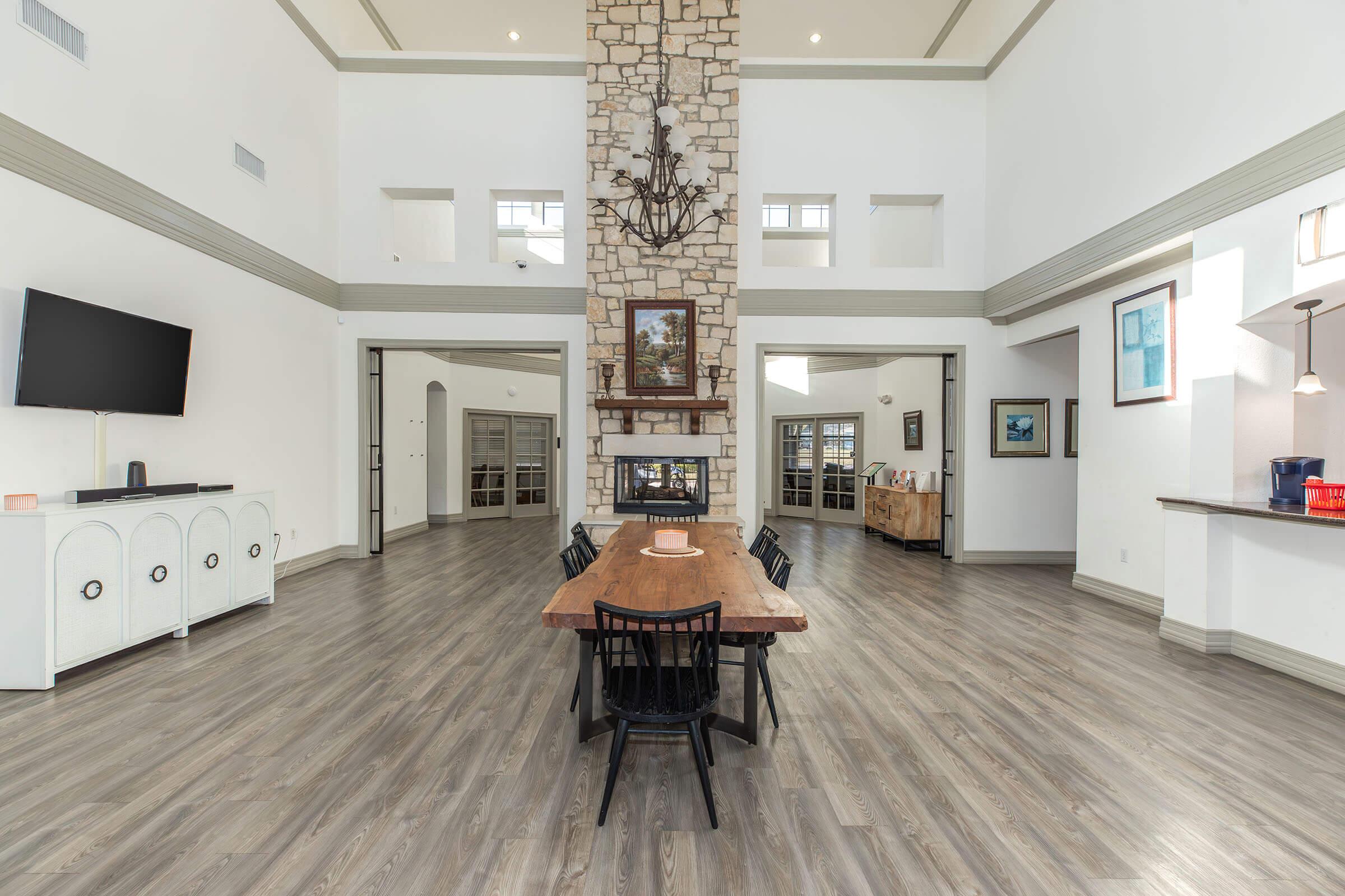 A spacious and modern interior featuring a large wooden dining table surrounded by black chairs. The room has high ceilings, a stone fireplace with a mantle, and large windows allowing natural light. A television is mounted on the wall, and an open layout connects to other areas of the home.