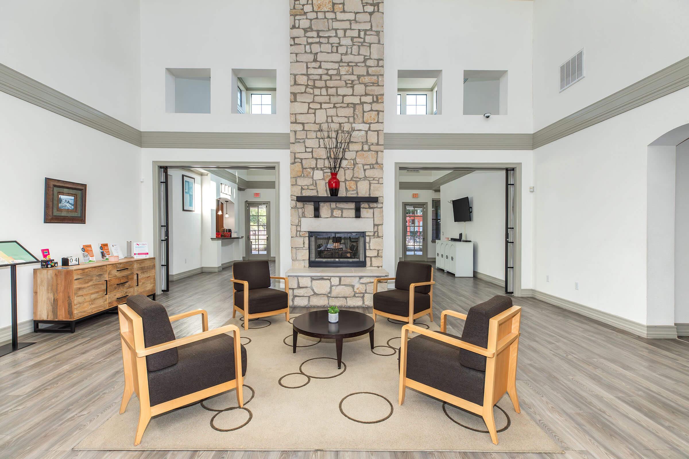 A spacious common area featuring a stone fireplace at the center, surrounded by four armchairs and a circular coffee table. Large windows provide natural light, and there is a wooden side table displaying brochures. The walls are painted in light colors, creating a modern and inviting atmosphere.