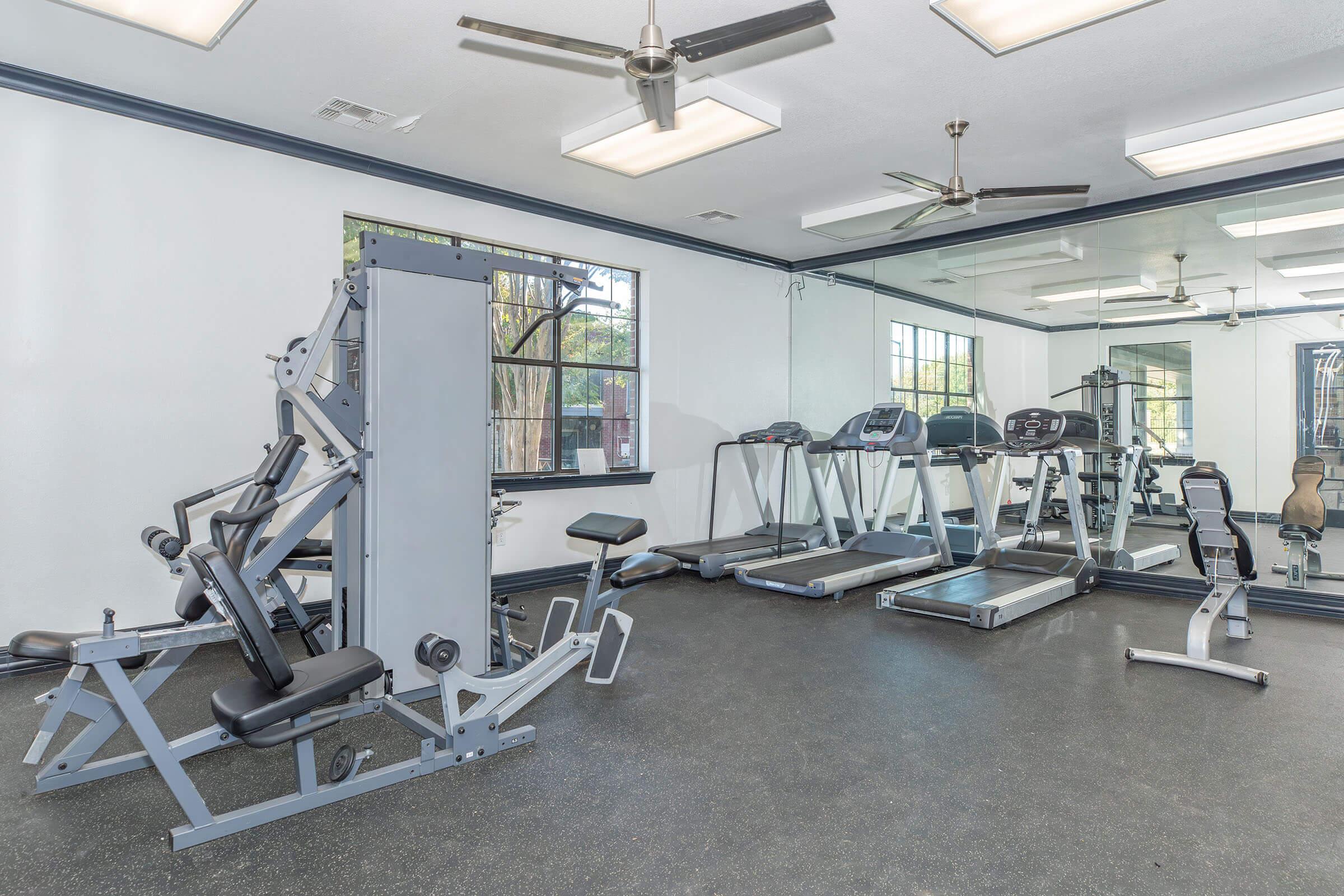 A modern gym interior featuring various exercise equipment, including weight machines, treadmills, and a stationary bike. The space is well-lit with large windows and mirrors, and has a rubber flooring for safety. Ceiling fans are present for ventilation.
