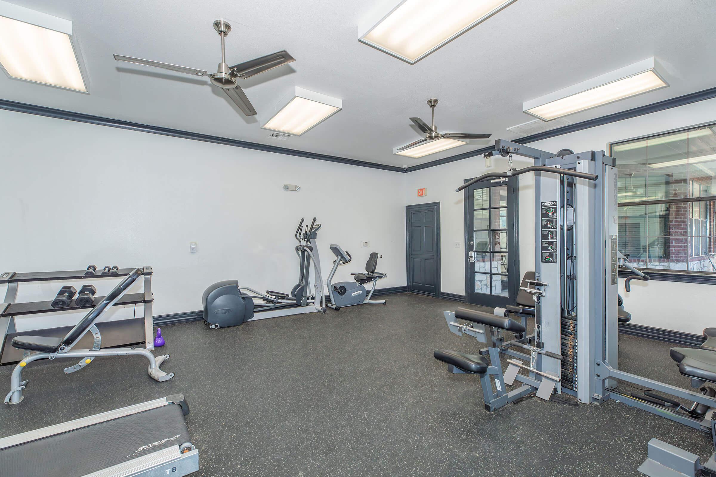 A bright gym interior featuring various workout equipment, including free weights, an elliptical machine, and a multi-station weight machine. The space is well-lit with ceiling fans and has a clean, open layout, designed for fitness activities.