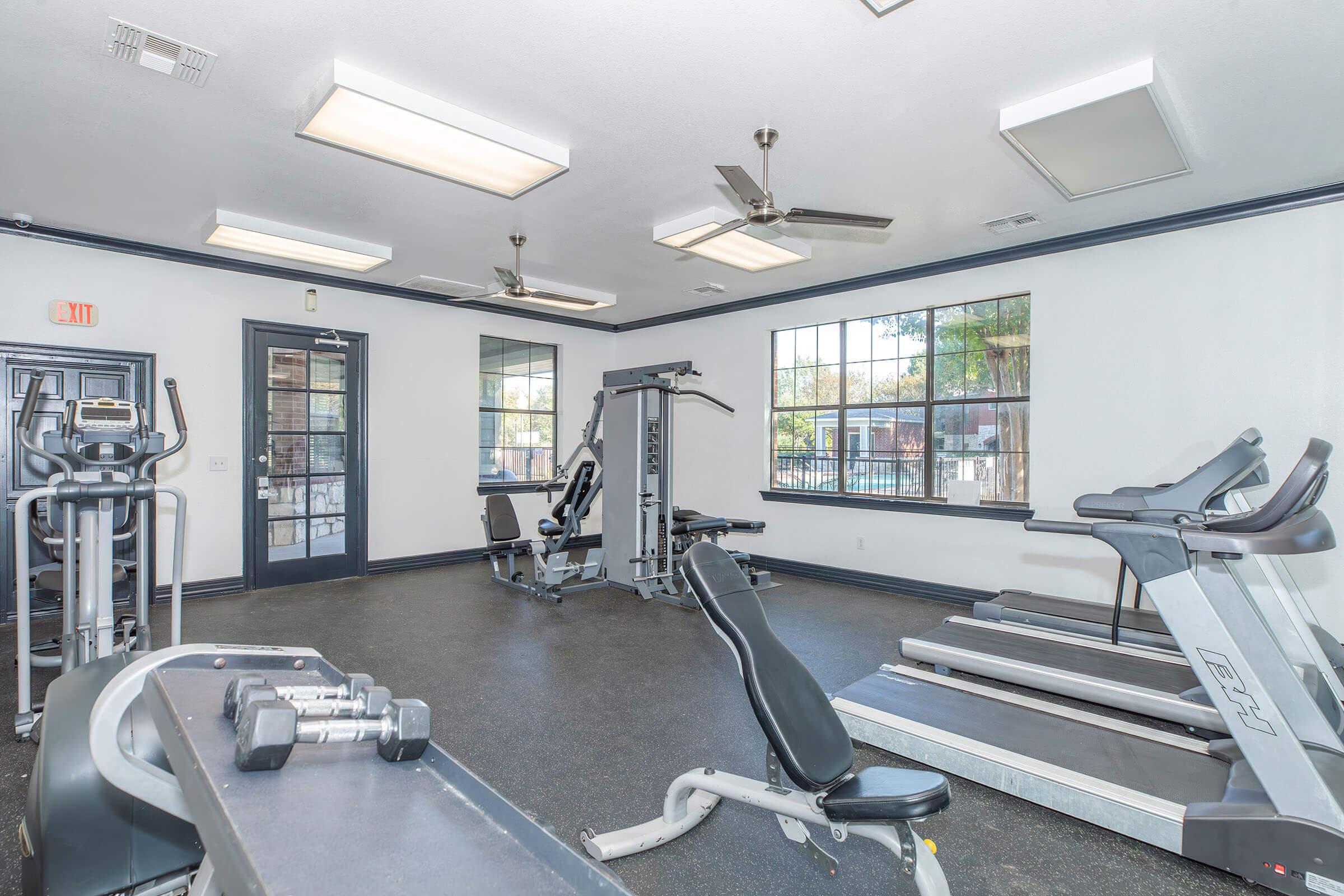 A modern fitness center featuring various gym equipment, including treadmills, an elliptical trainer, and strength training machines. The space is well-lit with large windows and ceiling fans, creating a bright and inviting atmosphere for workouts.