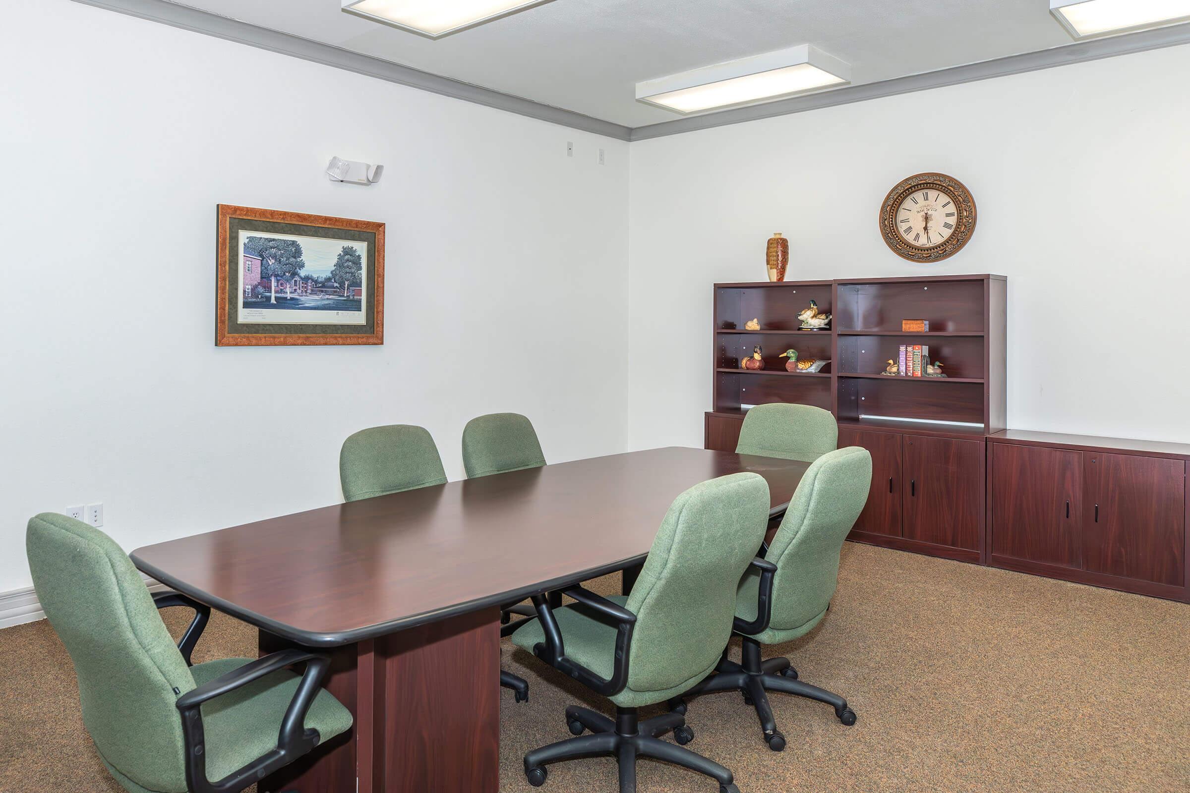 A sparsely furnished conference room featuring a large wooden table surrounded by green chairs. A framed landscape painting hangs on the white wall, and a shelf with decorative items and a clock is visible in the background. The floor is carpeted, and the lighting is bright and even.