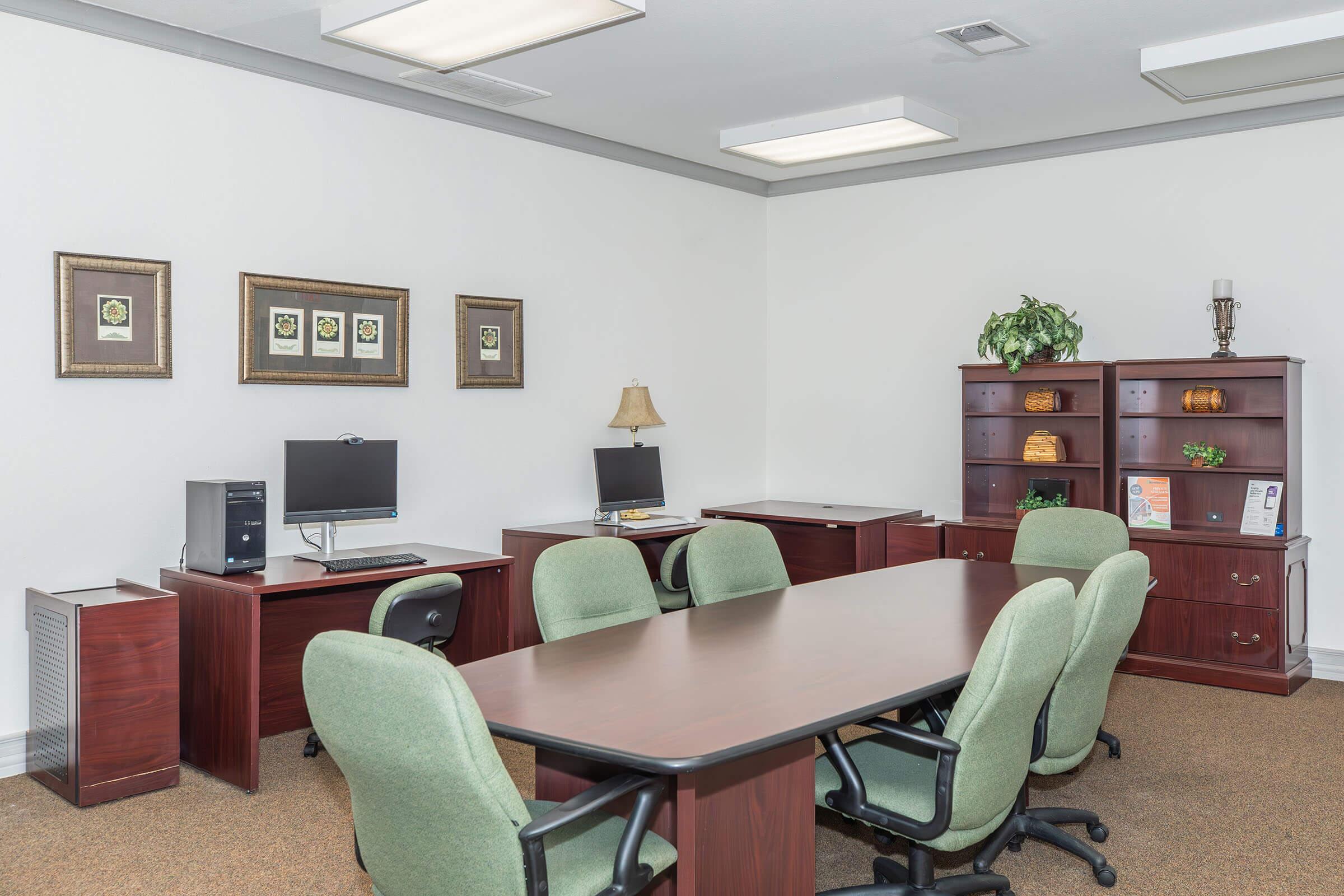 A modern office space featuring a large wooden table surrounded by green chairs. There are two computers on desks, a bookshelf with decorative items, and framed artwork on the walls. The floor is carpeted, and there is a potted plant in the corner, creating a professional and inviting atmosphere.