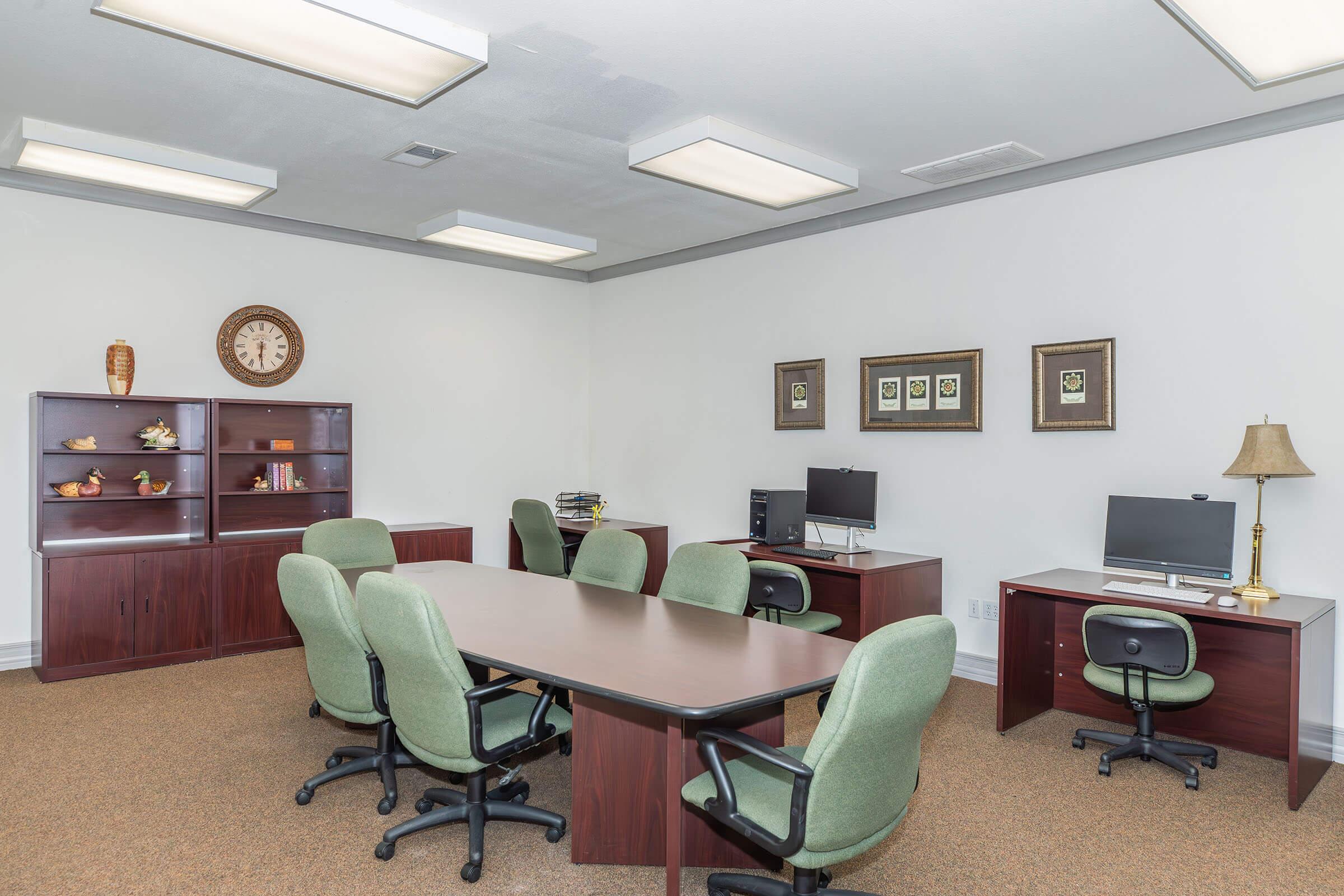 A modern office space featuring a large meeting table surrounded by green chairs, two computers on desks, a bookshelf with decorative items, a wall clock, and framed artwork. The walls are painted white, and the flooring is carpeted in a neutral tone. Bright overhead lights illuminate the room.