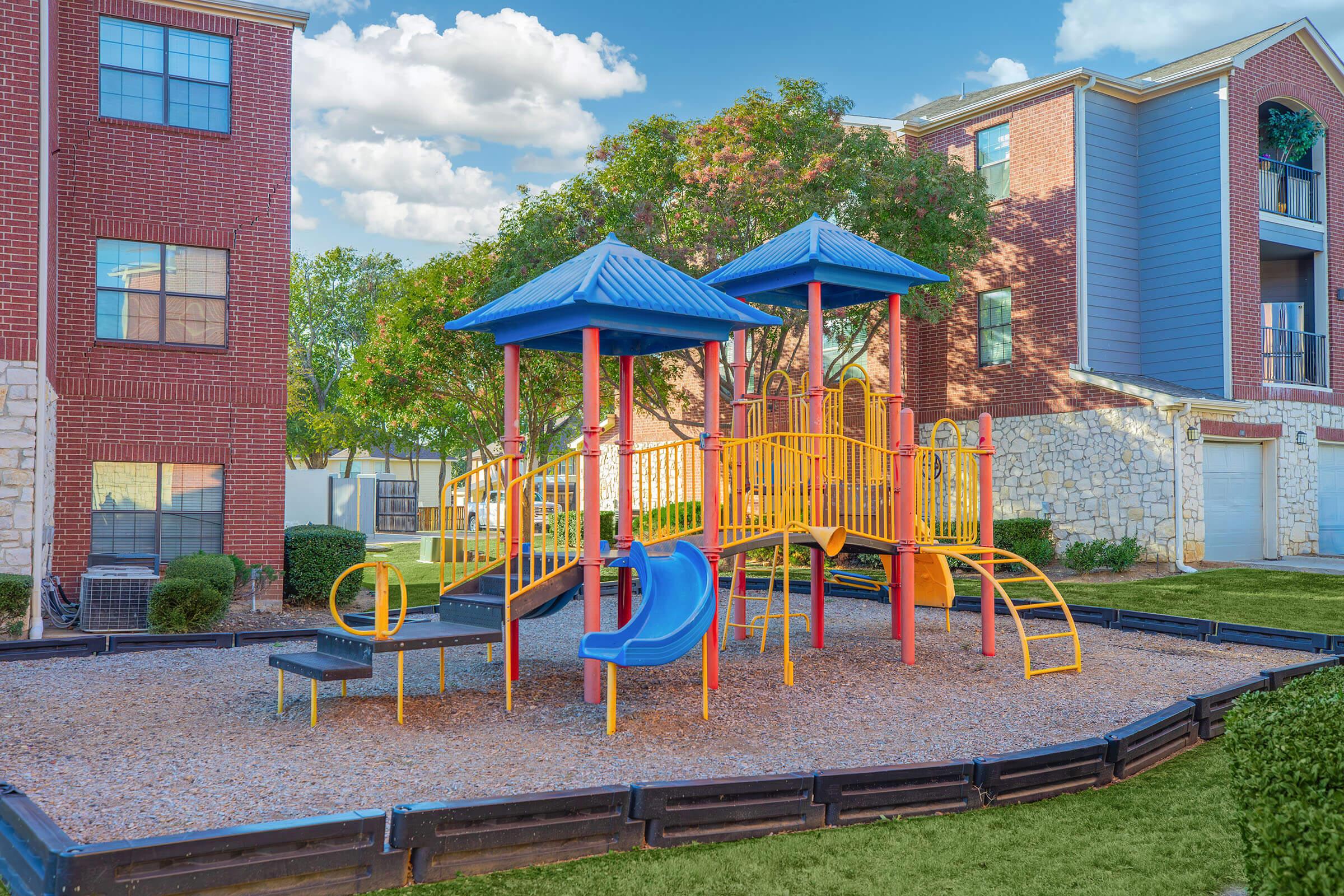 Colorful children's playground featuring a yellow and blue play structure with slides and climbing features, surrounded by gravel. The playground is situated in a green area near residential buildings, with trees and well-maintained grass in the background at sunset.