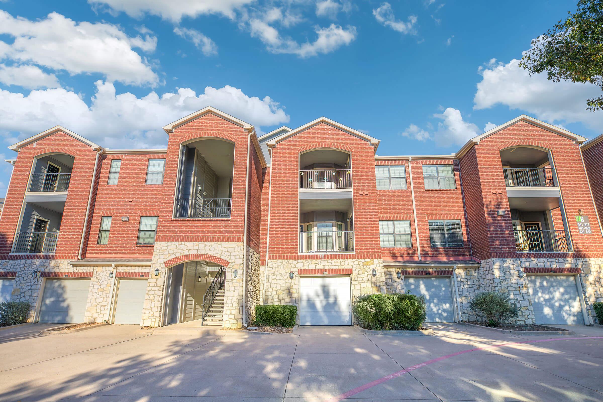 A two-story brick apartment building with a stone accent at the base. The building features multiple balconies, large windows, and garages beneath. The sky above is painted with soft pink and purple hues, suggesting either sunrise or sunset. Lush greenery surrounds the structure.