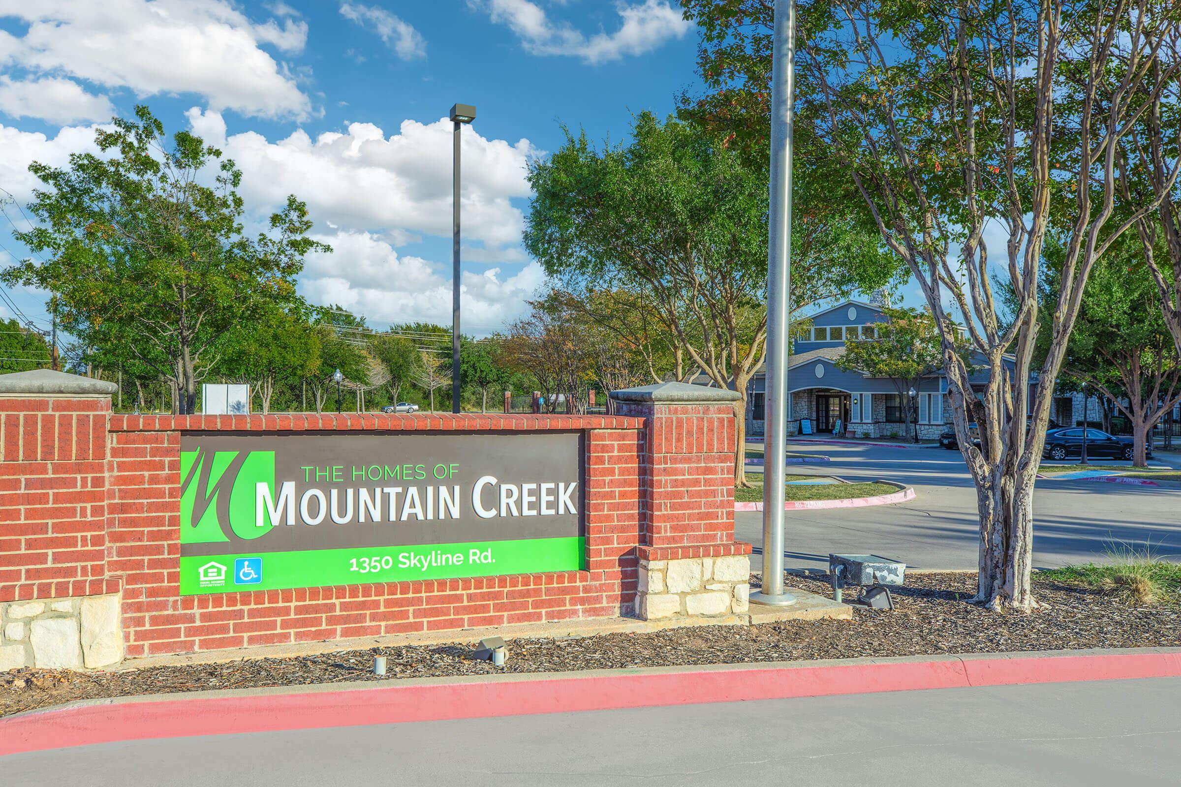 Sign for "The Homes of Mountain Creek" located at 1350 Skyline Rd, featuring a green and white logo against a red brick background, surrounded by trees and a clear sky.