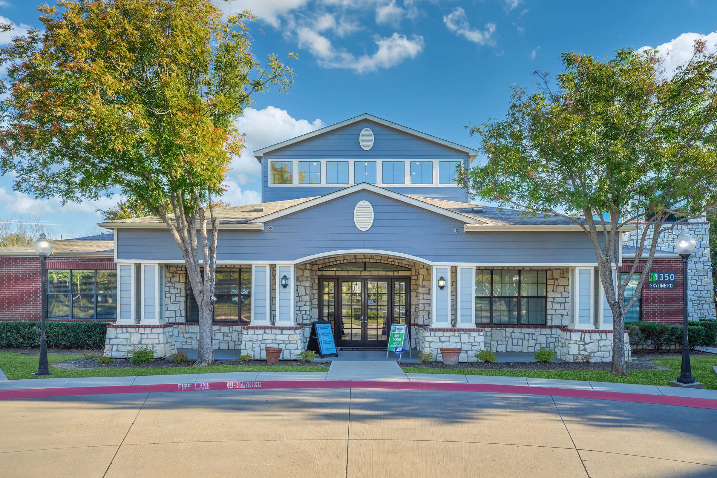 A modern building with a sloped roof and large windows, surrounded by trees and landscaped greenery. There are two signs in front, and the structure has a combination of stone and siding exterior. The sky features soft pink and blue hues, suggesting a sunset or sunrise.
