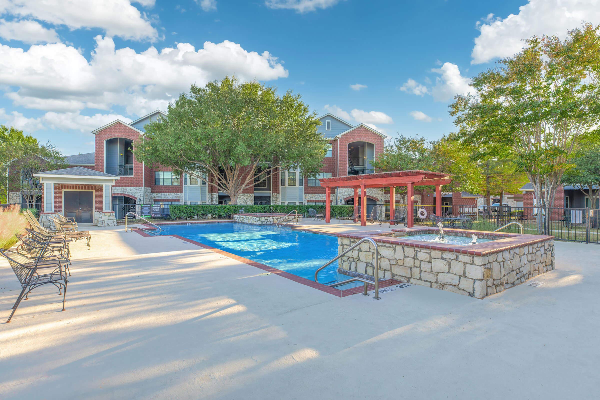 A swimming pool surrounded by a well-maintained area featuring lounge chairs and a stone border. There is a shaded pavilion nearby, and the background includes modern apartment buildings and lush trees, with a colorful sky at sunset.