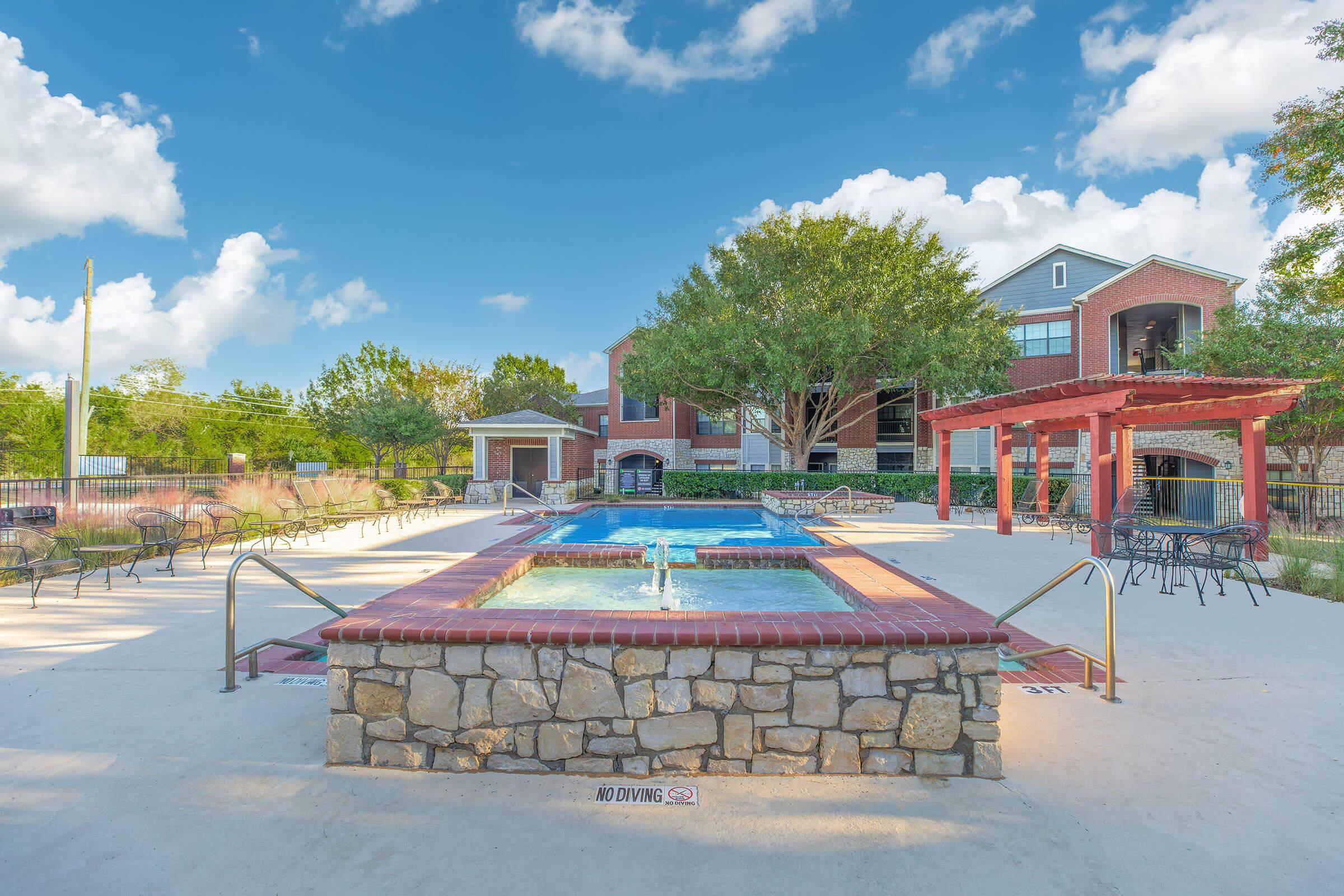 A well-maintained outdoor pool area featuring a large hot tub made of stone and a swimming pool, surrounded by lush trees and landscaped plants. In the background, there is a red pergola and a brick building, all set against a colorful sunset sky.