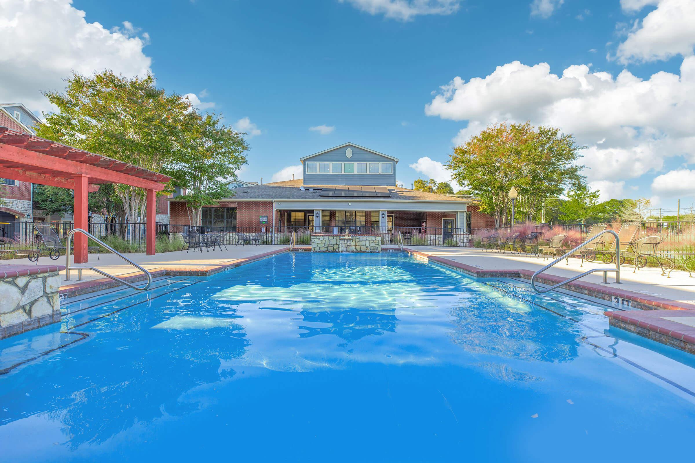 A serene swimming pool surrounded by a patio area with chairs and tables. In the background, a modern building with large windows is visible under a colorful sunset sky, featuring shades of pink and purple. Lush greenery frames the scene, creating a relaxing atmosphere.