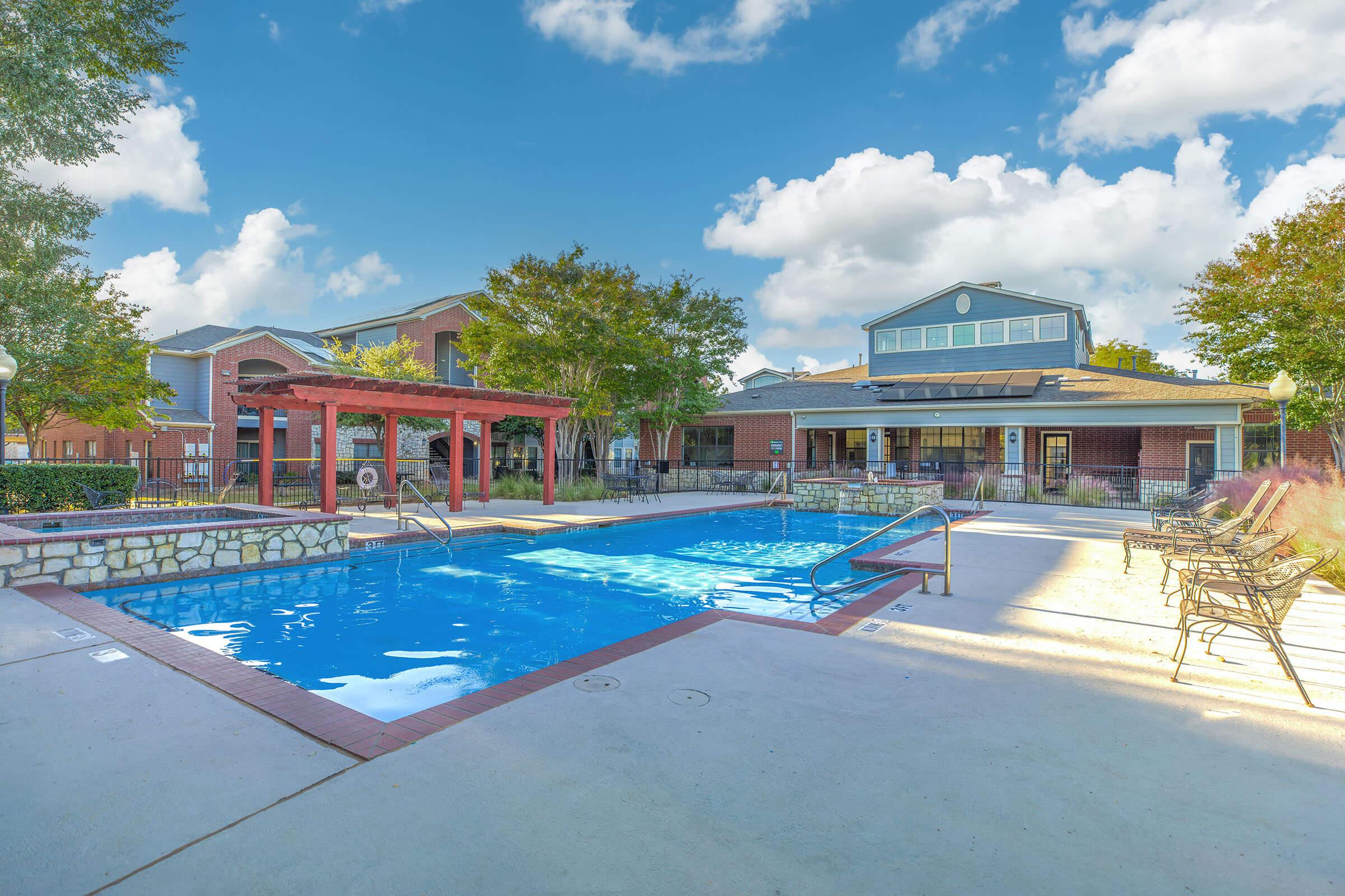 A tranquil pool area featuring a sparkling blue swimming pool, surrounded by lounge chairs and greenery. A pergola adds shade on one side, while a modern club house is visible in the background. The scene is illuminated by a beautiful sunset sky, enhancing the relaxing atmosphere.