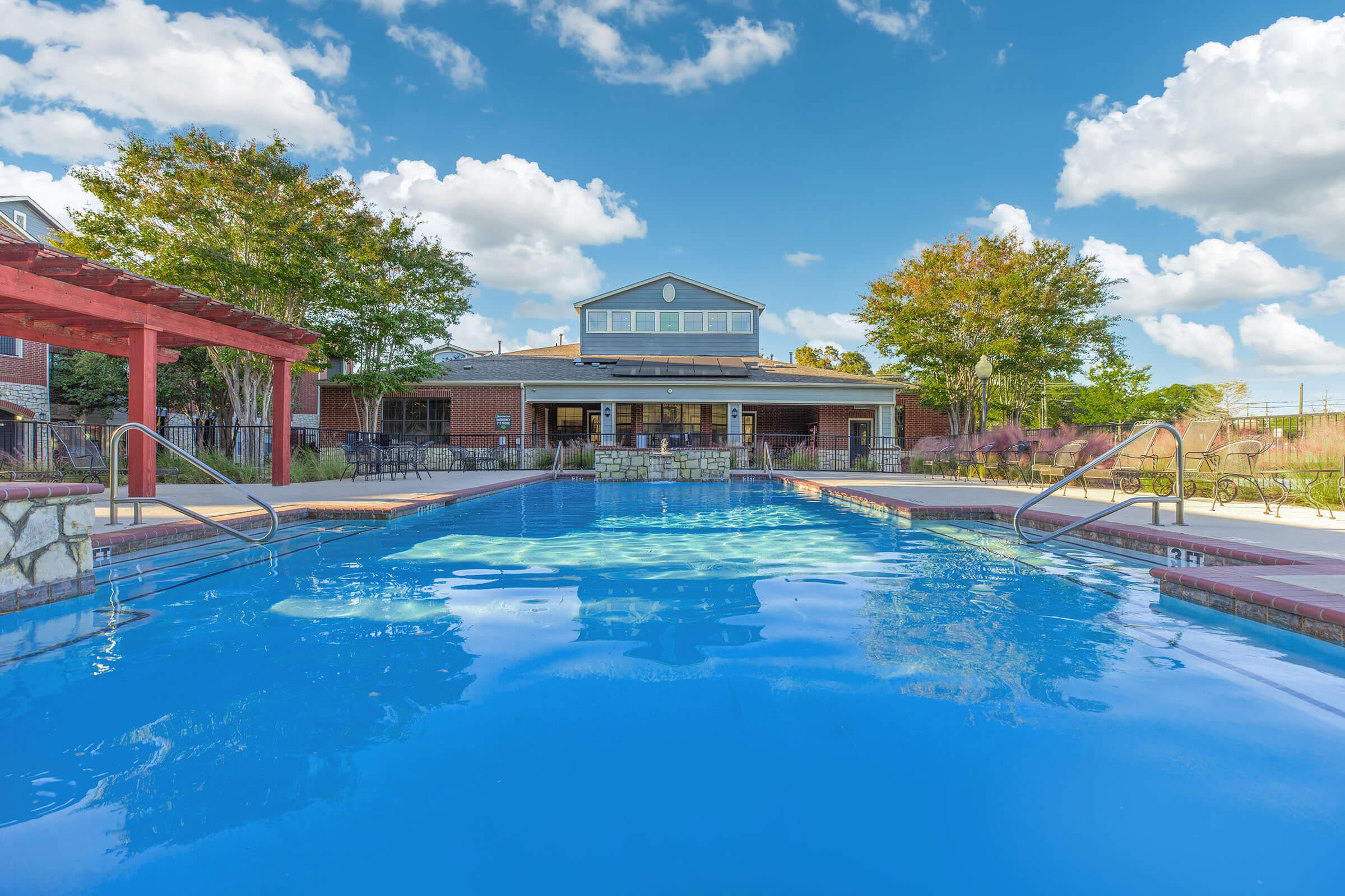 A serene outdoor swimming pool with clear blue water, surrounded by lush greenery and lounge chairs. In the background, a modern building with a large window overlooks the pool area, under a vibrant sunset sky filled with shades of pink and purple.