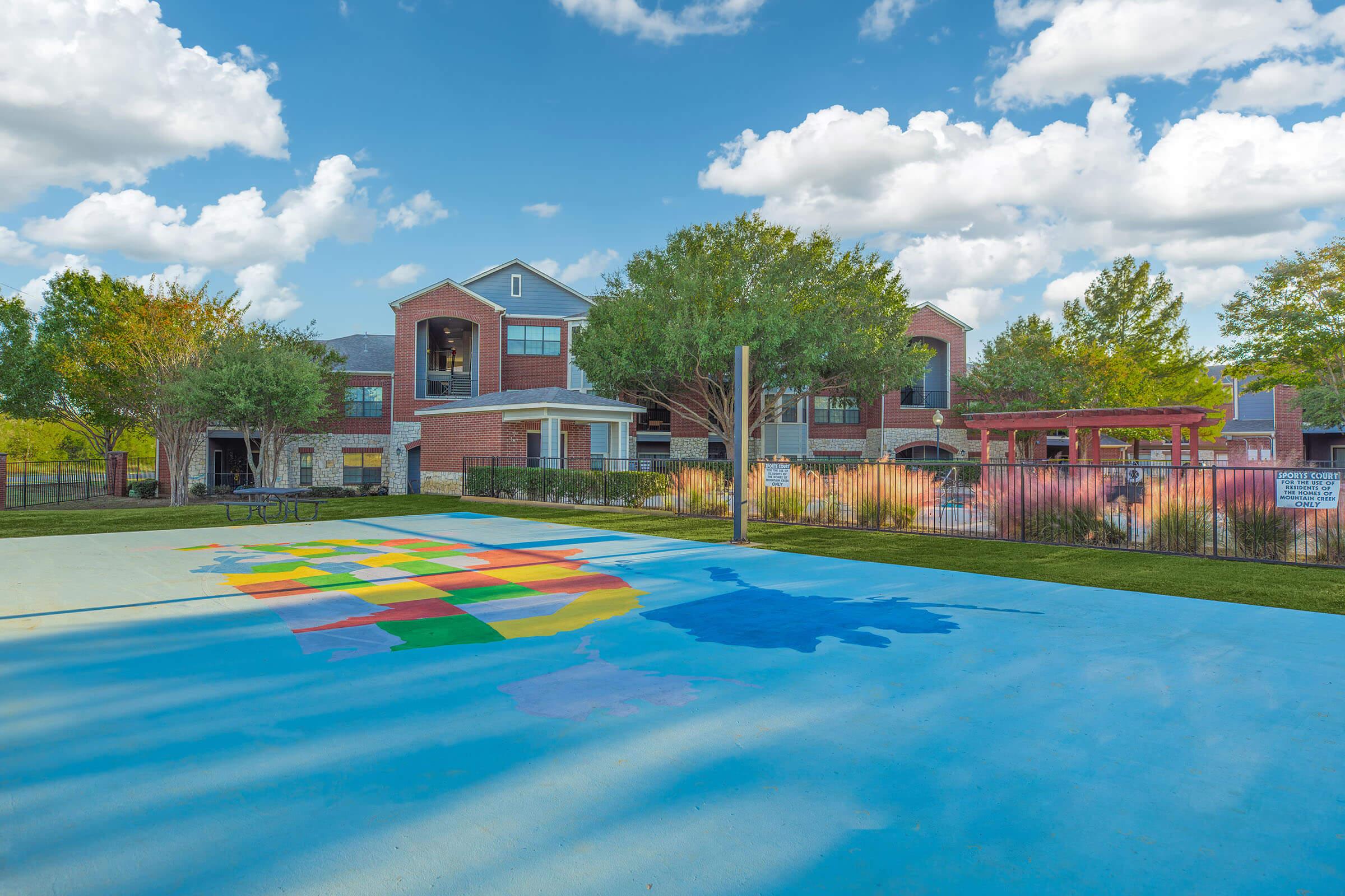 A colorful mural of a map on the ground, with various shades representing different regions. In the background, a modern building surrounded by trees and well-maintained landscaping. The sky features soft pastel colors during sunset. A playground area is visible near the mural.