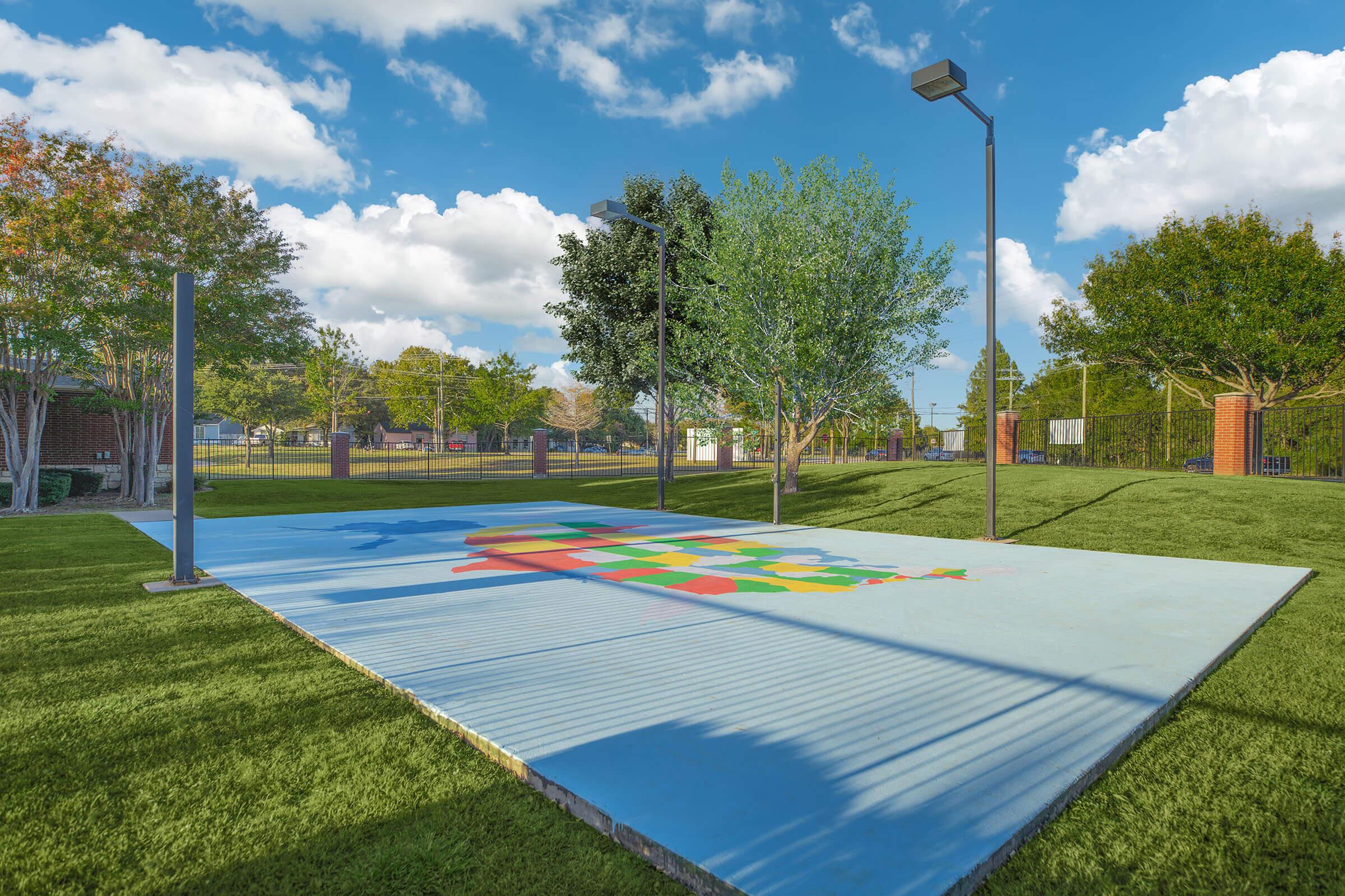 A colorful map of the United States is painted on a blue concrete surface in a grassy park. The scene features trees and lampposts under a pastel sky during sunset, creating a vibrant and inviting atmosphere. The park appears well-maintained with a spacious layout.