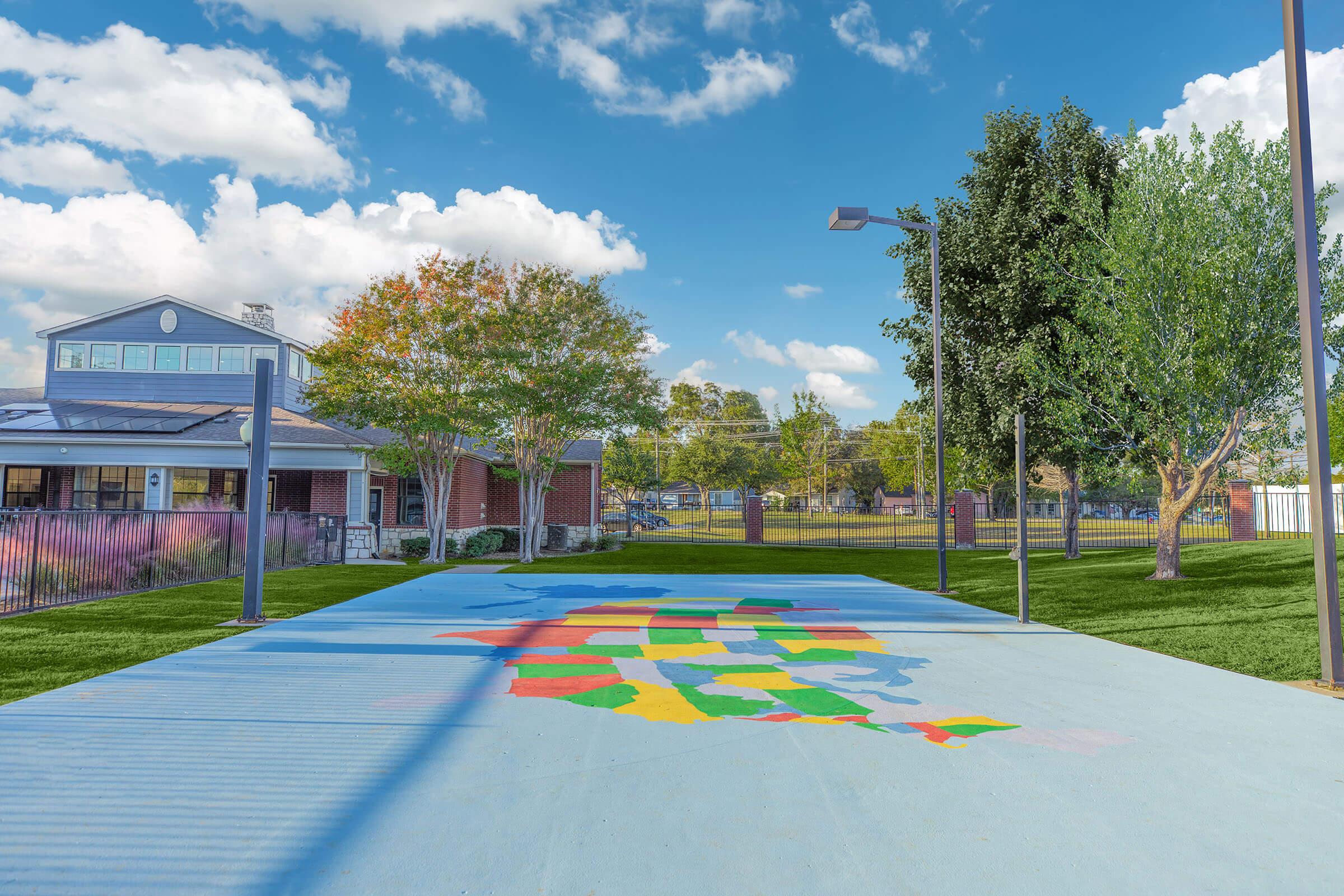 A vibrant mural of the United States map painted on a concrete playground surface, set against a colorful sunset sky. Surrounding the mural, there are trees and a modern building in the background, with scattered lighting fixtures illuminating the area. The scene conveys a cheerful outdoor space for recreation.