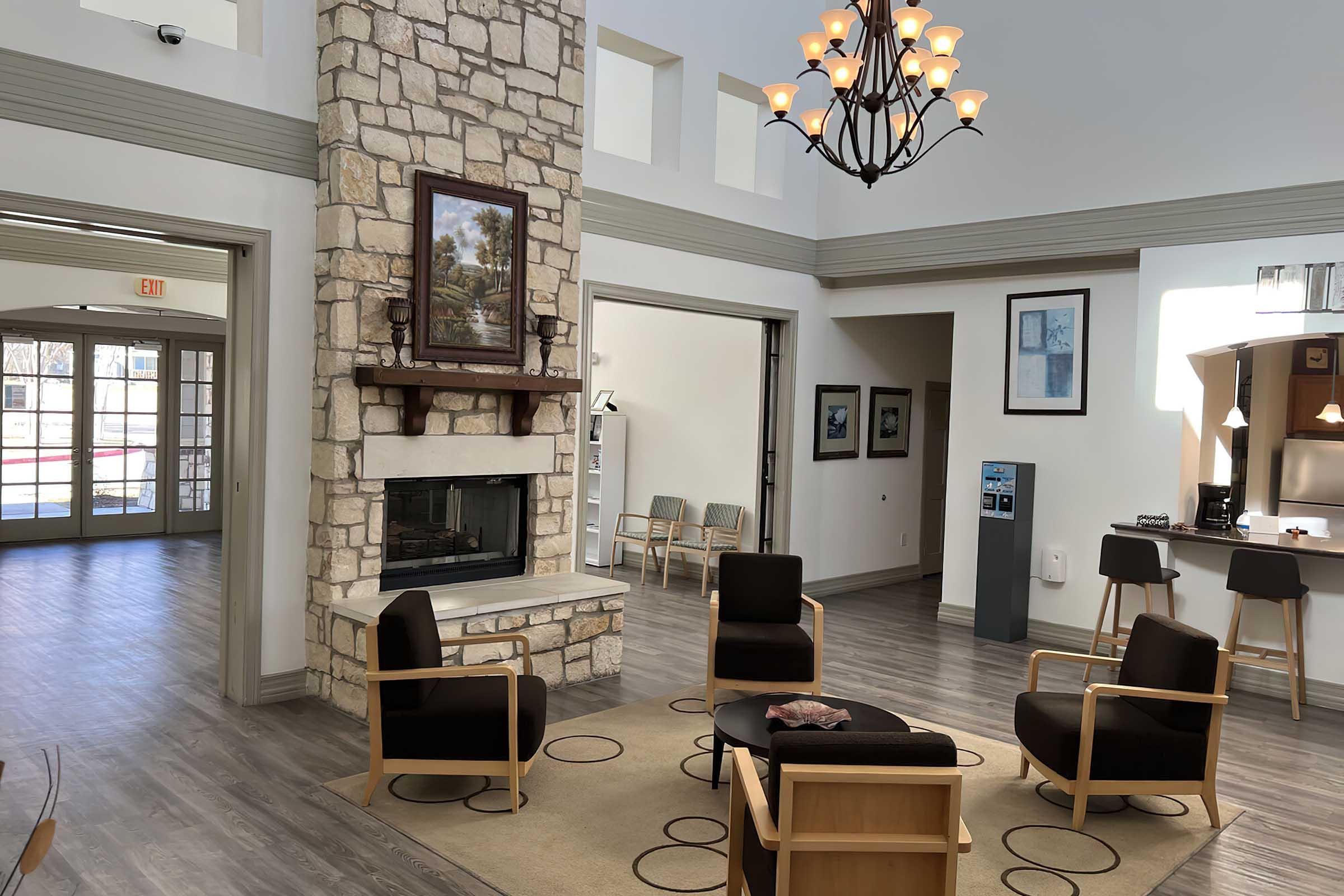 A spacious, modern interior of a community room featuring a stone fireplace, wooden chairs, and a chandelier.