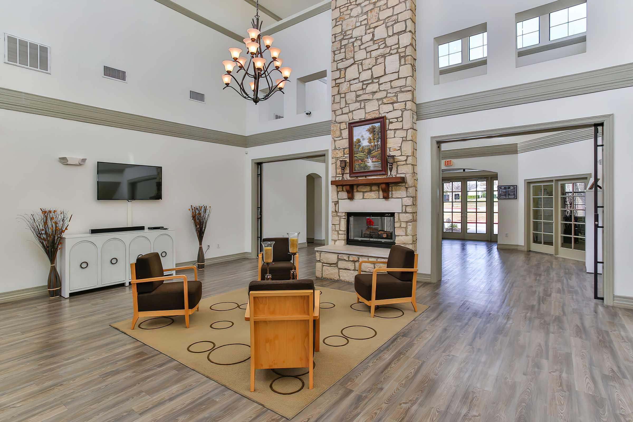A spacious, well-lit lounge area featuring a stone fireplace, comfortable seating with wooden chairs, a central area rug with circular patterns, and large windows