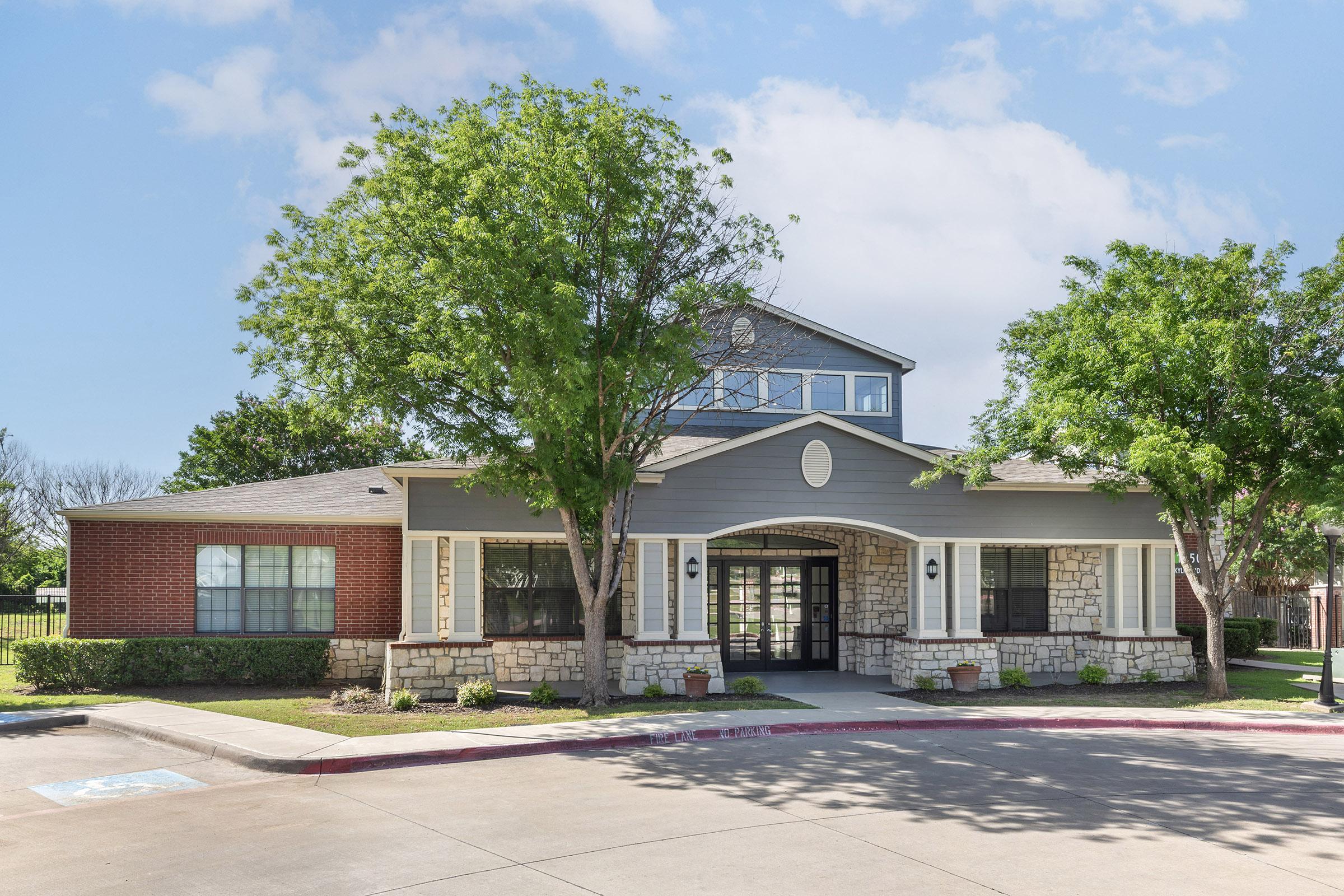 A modern building with a combination of stone and gray siding. It features large windows, a central entrance with double doors, and is surrounded by green trees and neatly landscaped bushes. The driveway is paved, and the sky is partly cloudy, creating a bright, inviting atmosphere.