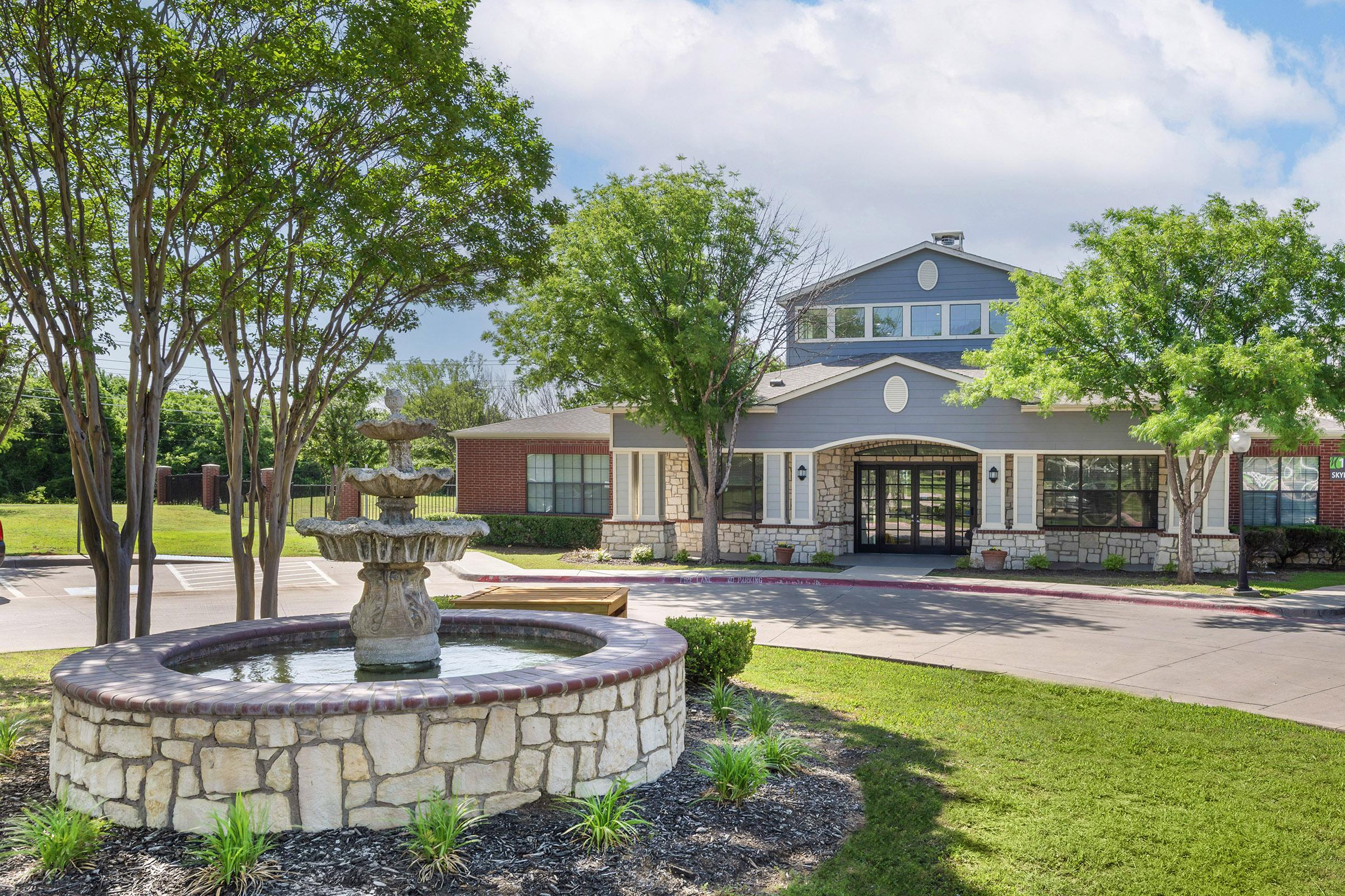 A landscaped exterior view of a building featuring a small fountain surrounded by greenery. The structure has a modern design with large windows and a welcoming entrance, set against a bright sky with scattered clouds.