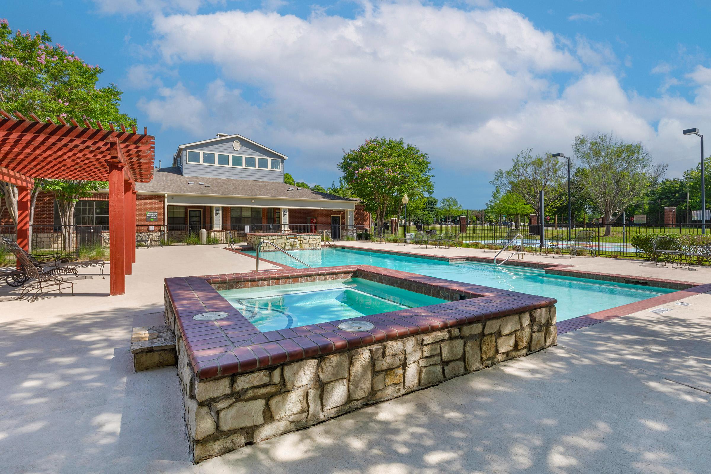 A sunny outdoor pool area featuring two swimming pools: one large and rectangular, and another smaller heated spa with stone edges. Surrounding the pools are lounge chairs, shaded by a pergola, and well-maintained greenery in the background. A clubhouse is visible in the distance under a blue sky with fluffy clouds.