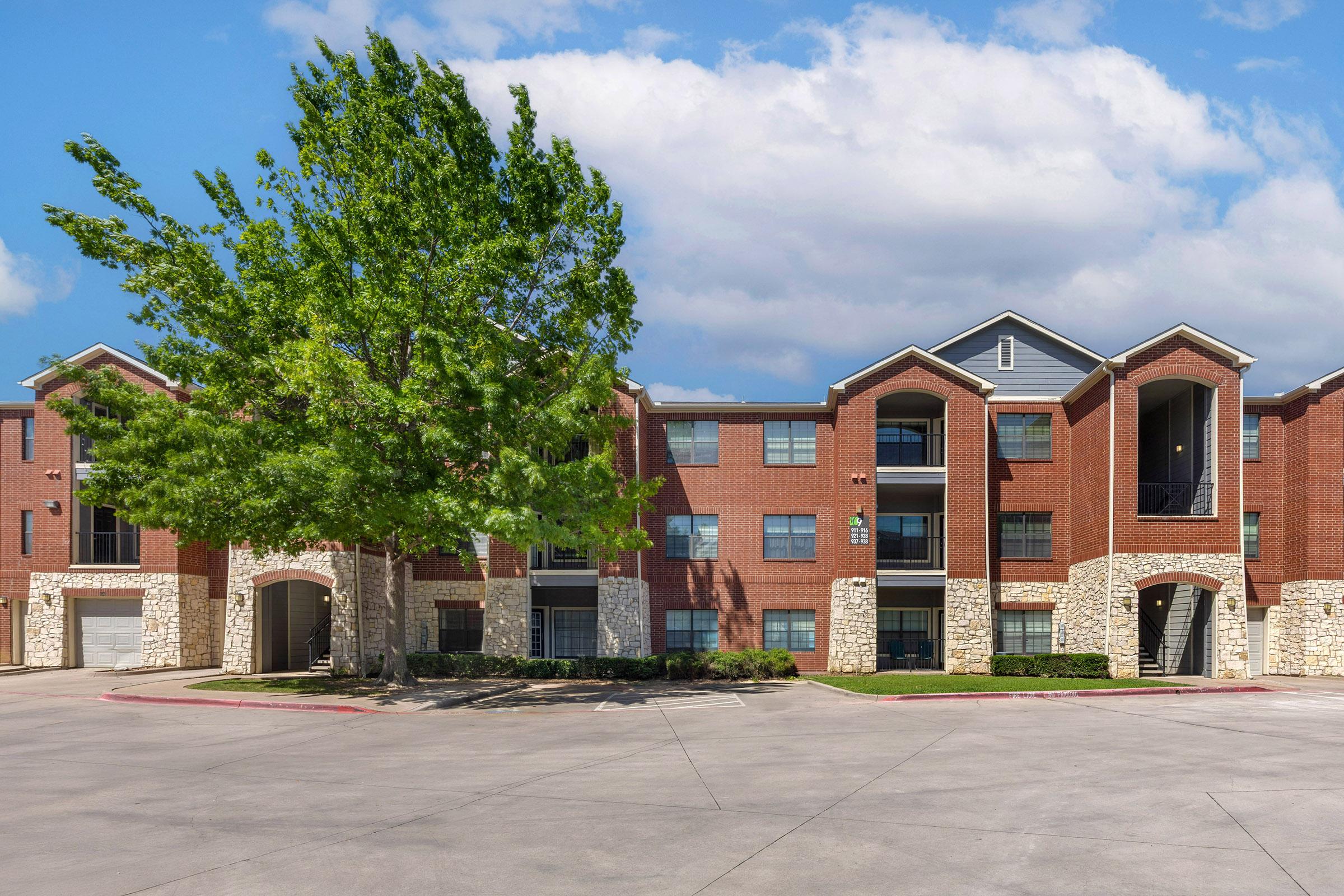 A residential apartment building with a brick facade and stone accents, featuring multiple windows and balconies. In front, there is a large green tree and paved parking space. The sky is partly cloudy, creating a bright and inviting atmosphere.