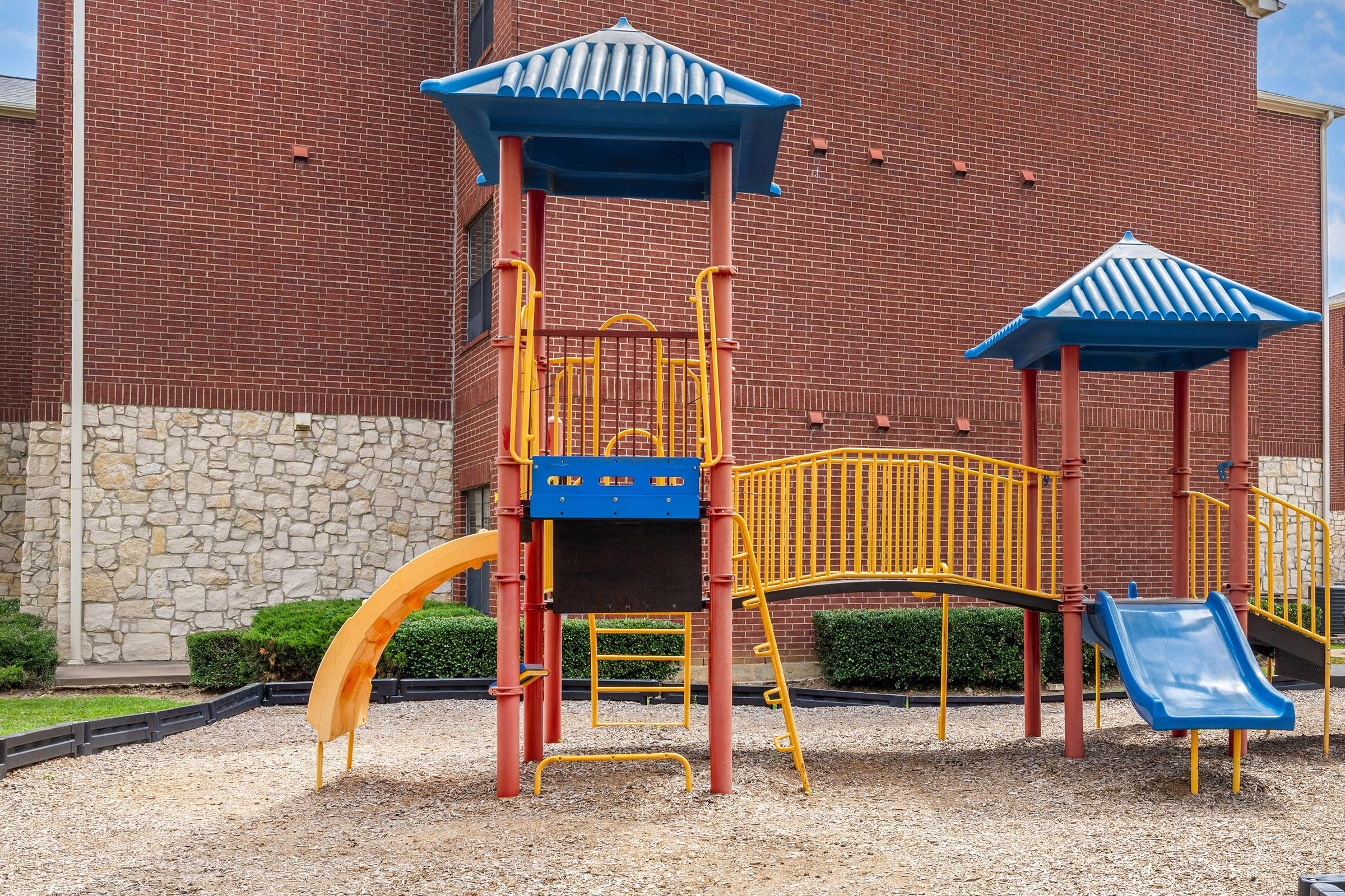 Colorful playground structure featuring blue and yellow slides, multiple climbing elements, and a roofed lookout tower, set on a gravel surface with green bushes in the background and a brick wall behind.