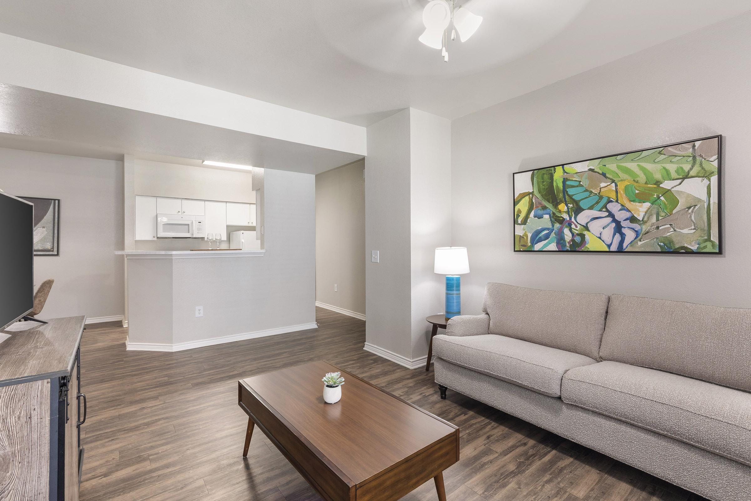 A modern living room featuring a light gray sofa, a wooden coffee table with a small plant, and a wall-mounted TV. The space has warm wood flooring and soft lighting. A colorful abstract painting hangs above the sofa, and the open kitchen area is visible in the background with white cabinets and appliances.
