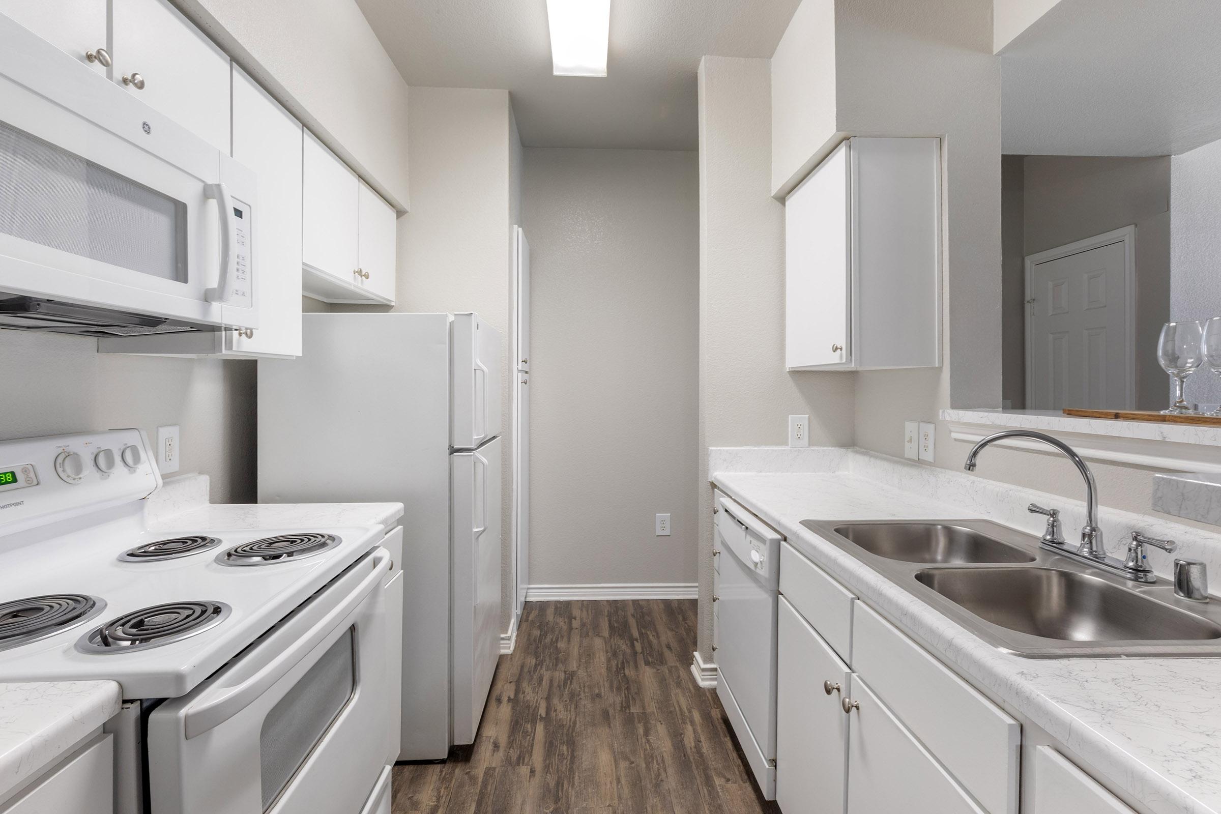 A modern kitchen featuring white cabinets, a double sink, and stainless steel appliances, including a stove, microwave, and refrigerator. The countertops are light-colored with a marble-like finish, and the flooring is dark wood laminate, creating a contrasting warm atmosphere.