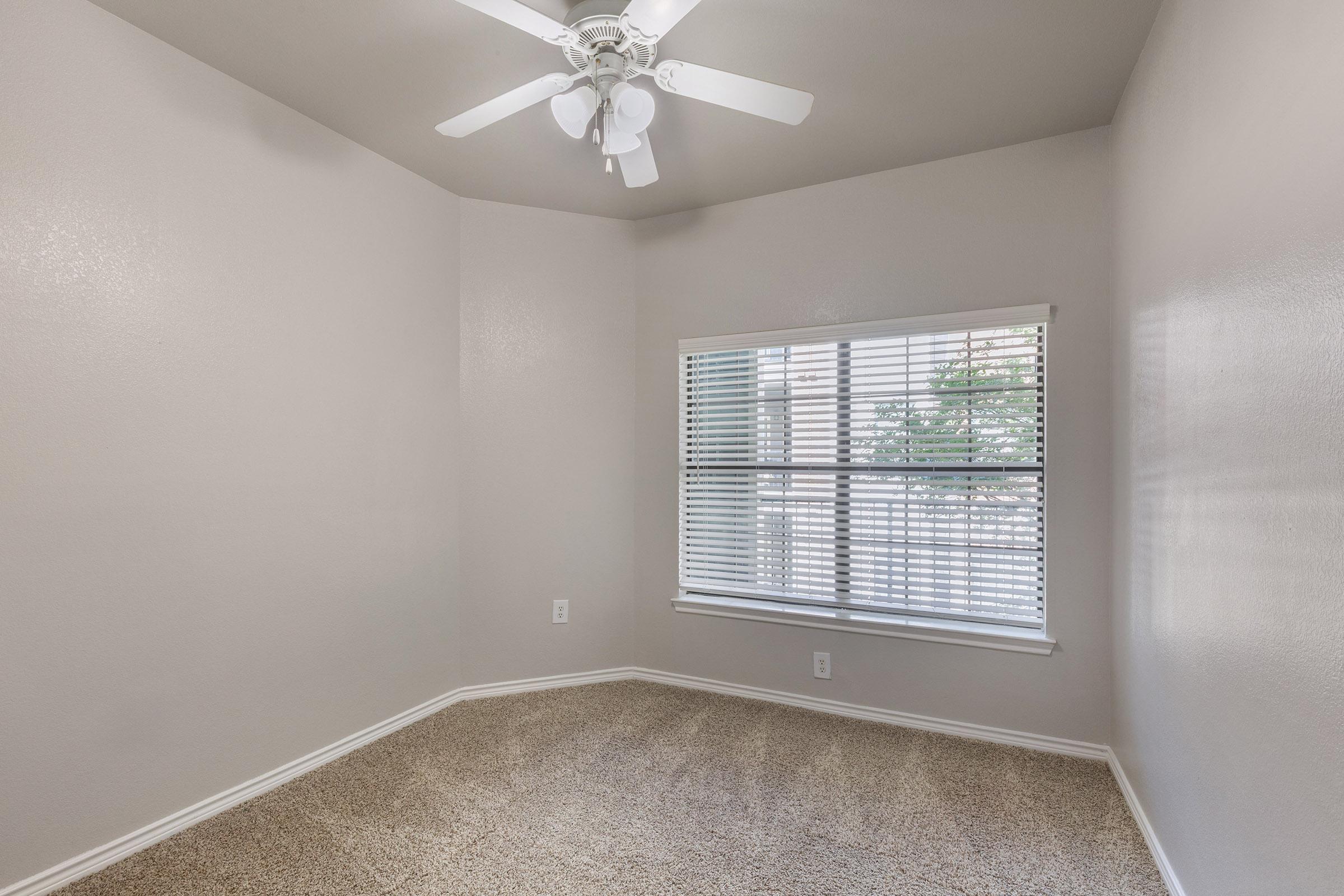 A small, empty room featuring light beige walls, a ceiling fan with white blades, and a large window adorned with blinds. The floor is covered with carpet in a similar beige tone, and there are no furnishings present, giving the space a clean, minimalistic appearance.