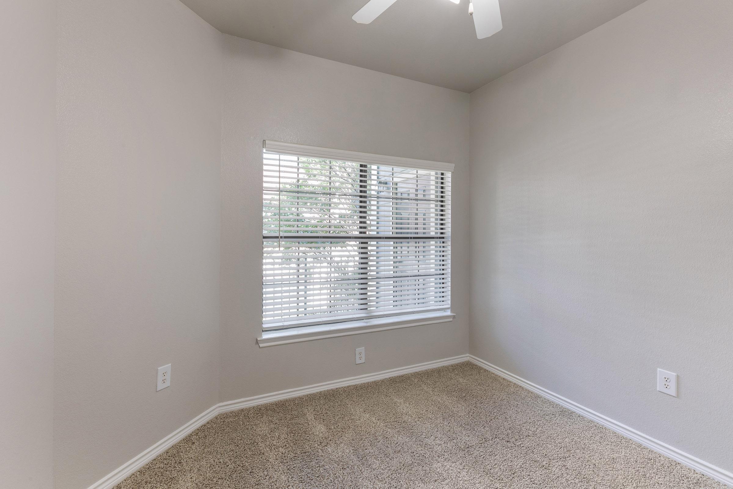 A small, empty room with light gray walls and beige carpeting. There's a large window with white blinds allowing natural light to enter. The room has a ceiling fan and minimal decor, creating a clean and simple aesthetic.