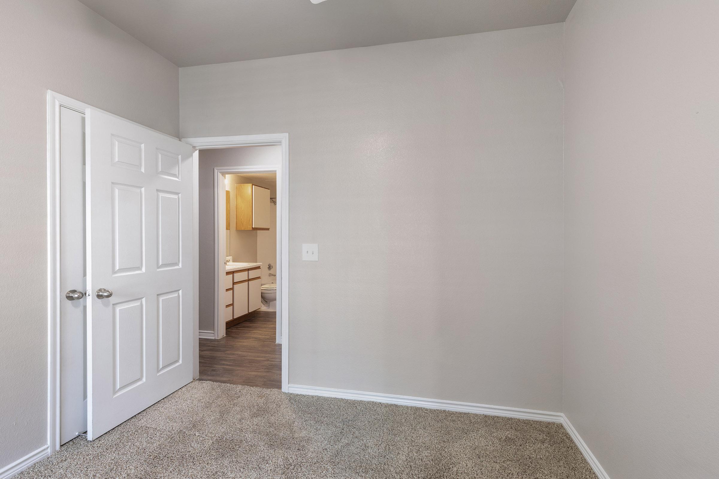 A view of a small, empty room with beige walls and carpeted flooring. A door is open, leading to a bathroom area with light wooden cabinets. Natural light enters through the doorway, creating a bright and airy atmosphere in the space. The room has no furniture, emphasizing its emptiness.