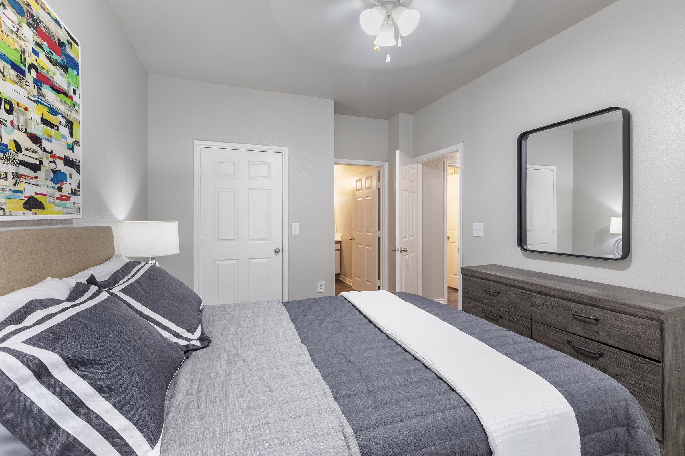 A modern bedroom featuring a queen-sized bed with gray and white bedding, a large mirror above a wooden dresser, a colorful abstract painting on the wall, and doors leading to adjacent rooms. Ceiling fan and neutral wall colors create a cozy atmosphere.