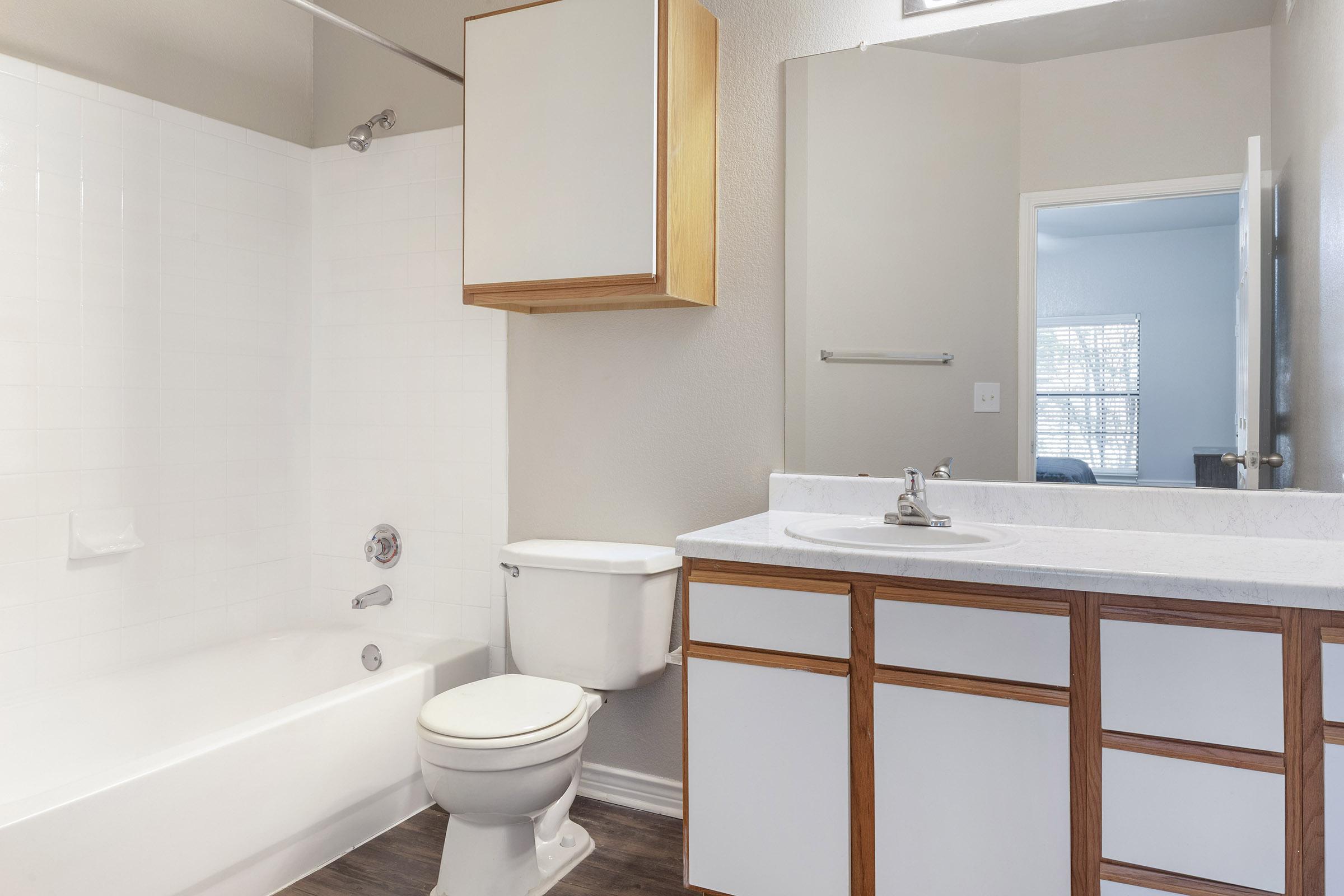 A clean and modern bathroom featuring a white bathtub, a toilet, and a vanity with a sink. The walls are painted light beige, and a wooden storage cabinet is mounted above the sink. A mirror reflects part of the room, which has a door leading to an outside area and a window allowing natural light.