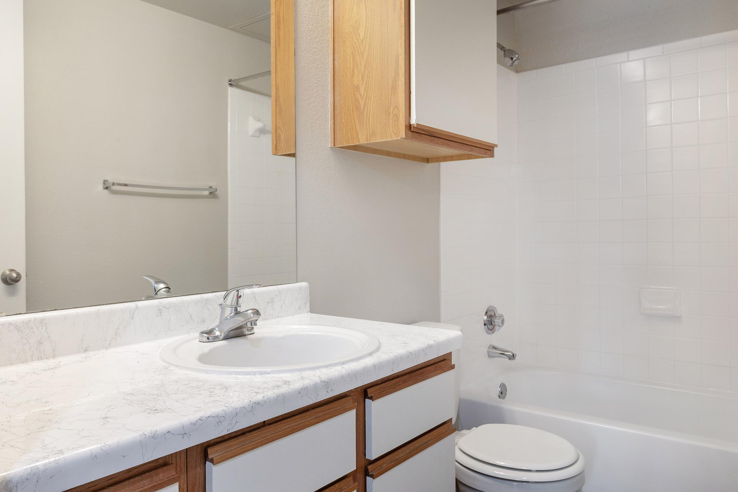 A clean and modern bathroom featuring a white bathtub, a sink with a marble countertop, wooden cabinets, and a large mirror. The walls are light-colored, and there's a towel bar on the wall. The space appears well-lit and orderly, highlighting a minimalist design.