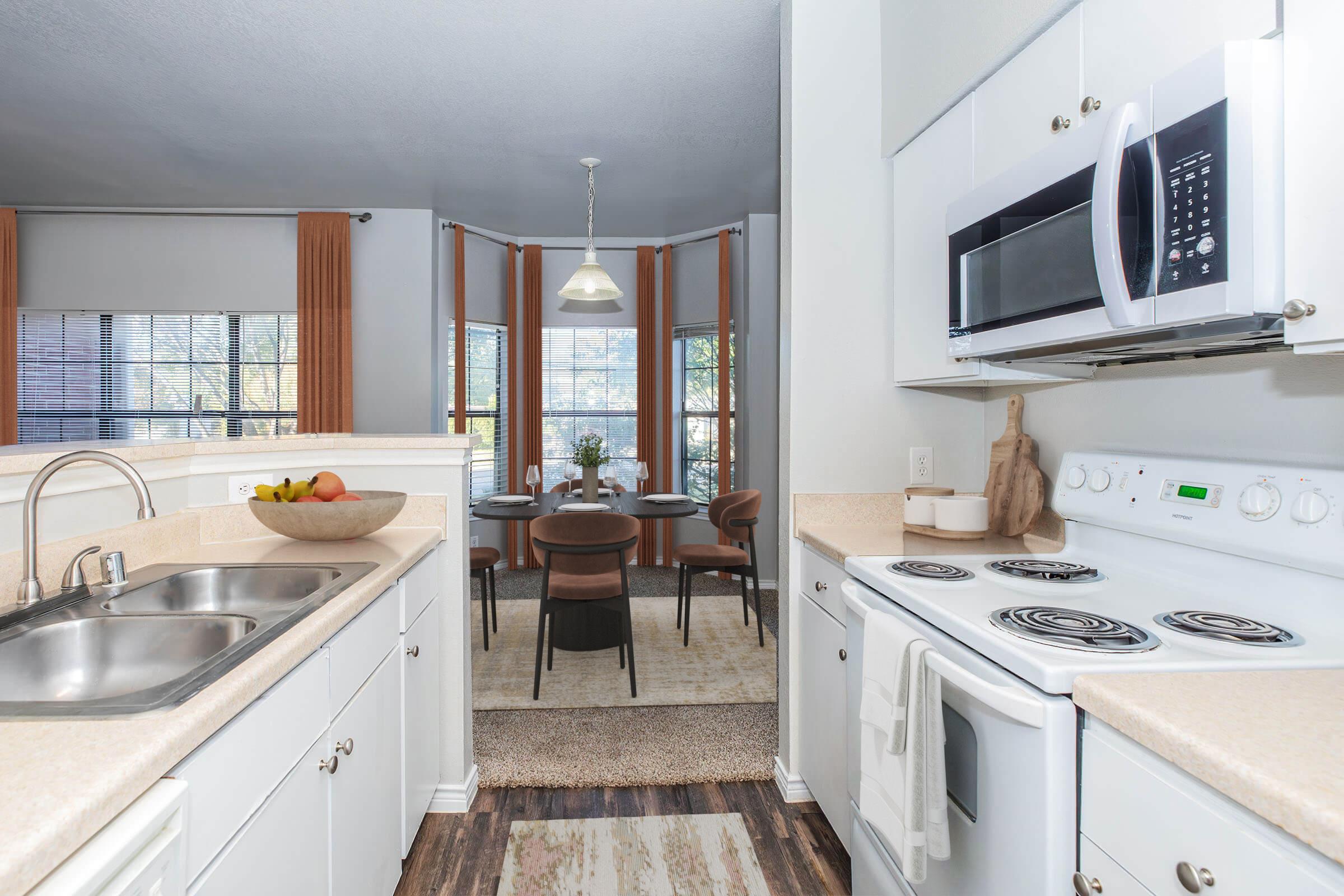 Bright and modern kitchen featuring a double sink, stove, and microwave. The kitchen overlooks a dining area with a round table and chairs, illuminated by a pendant light. Large windows bring in natural light, complemented by neutral-toned decor and a cozy atmosphere.