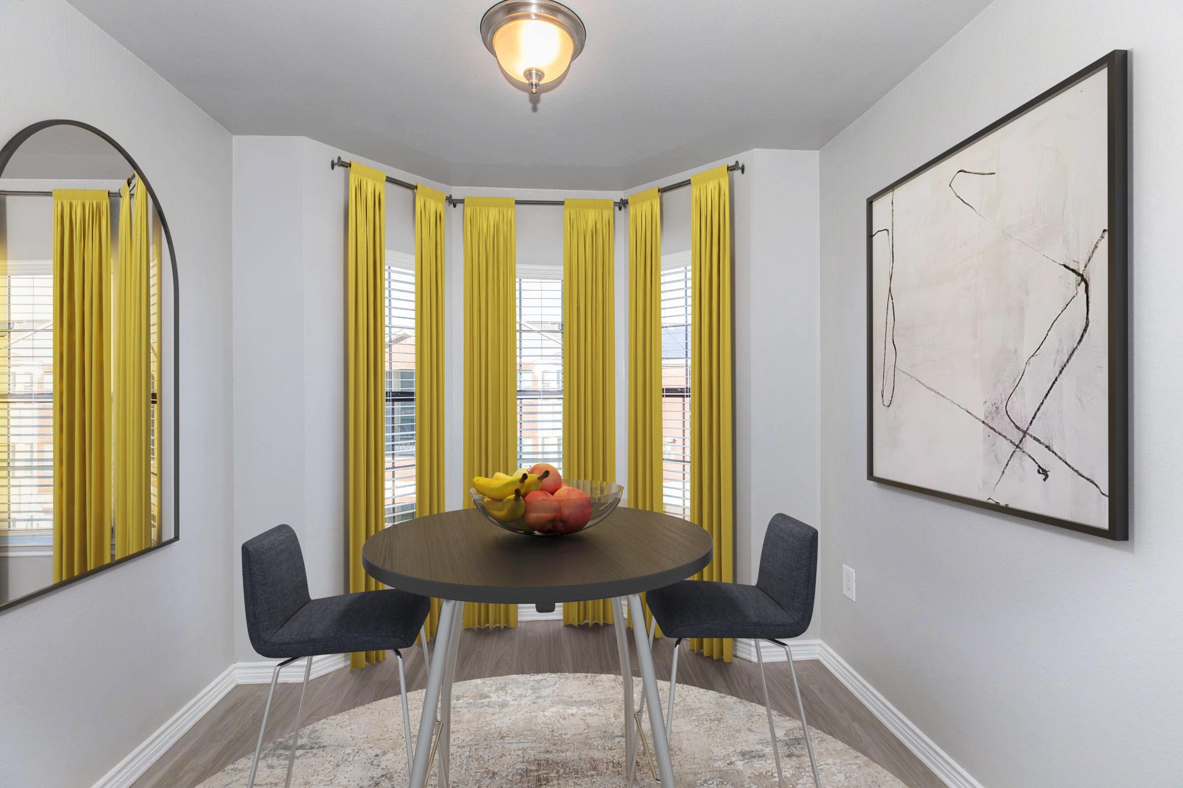 A cozy dining nook featuring a round table with a fruit bowl in the center, surrounded by two modern chairs. Large windows with yellow curtains allow natural light to fill the space. A circular mirror is mounted on the wall, and an abstract painting adds a touch of style to the room.