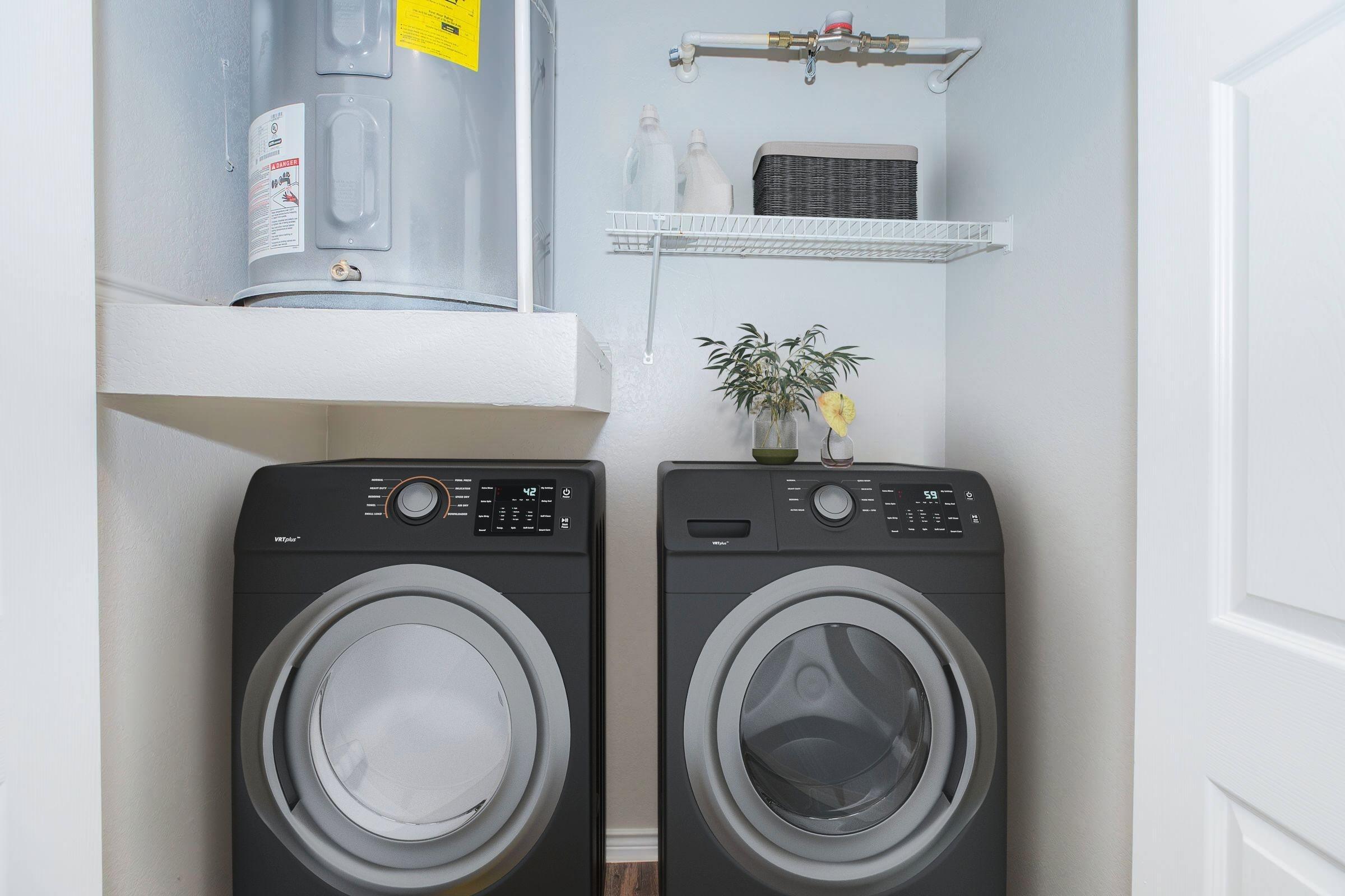A laundry room featuring a black washing machine and dryer, stacked neatly beside each other. Above them, there is a shelf with a small plant and a basket. A water heater is visible on the wall, and the space is well-organized and clean.