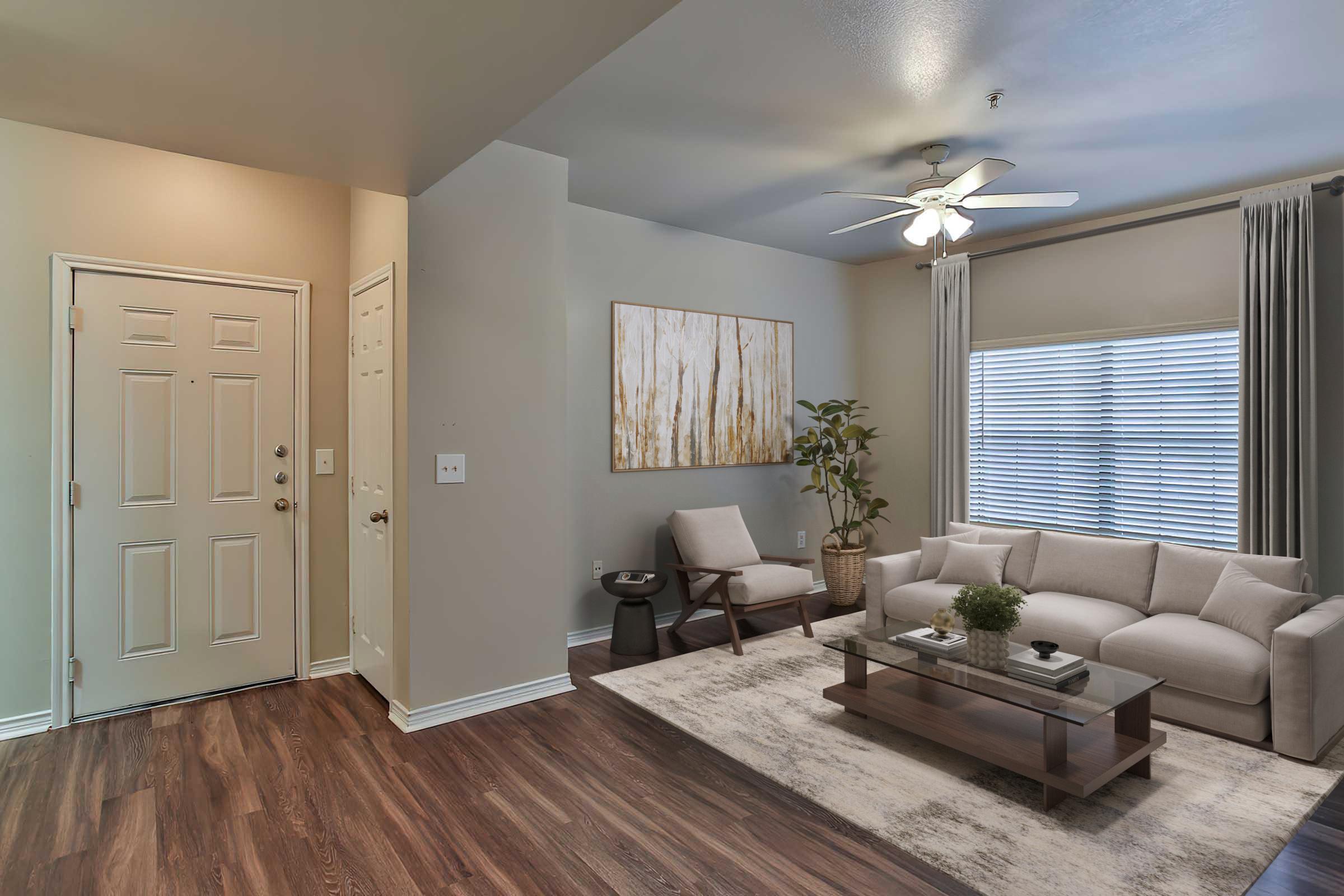 A modern living room featuring a light-colored sofa, a glass coffee table with a small plant, and a wooden floor. There’s a wall art piece with abstract designs, a ceiling fan, and large windows dressed with curtains. The entrance door and a chair are visible in the corner of the room.